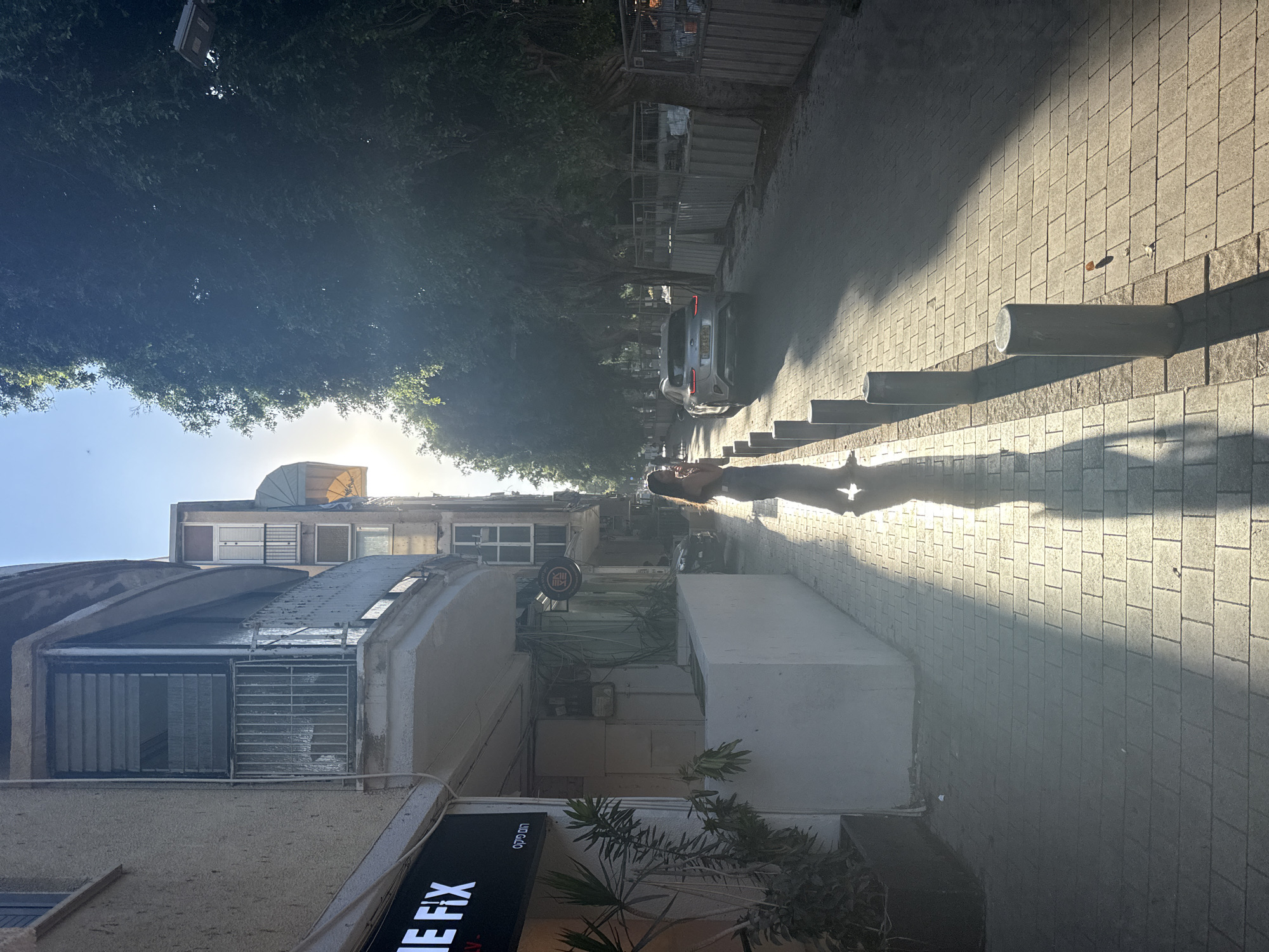 This image depicts a street scene in Tel Aviv, Israel, likely during late afternoon or early morning due to the low angle of the sun casting long shadows. A young woman with dark hair, wearing a dark top and wide-legged pants, stands on a paved sidewalk, her silhouette clearly defined by the strong sunlight. She appears to be holding a phone to her ear, engaged in a conversation.
The sidewalk is lined with bollards, and a light-colored building with a balcony featuring vertical railings rises on the left. A business sign with white text on a black background, reading "IE FIX," is visible on this building. Further down the street, mature trees with dense foliage line the right side, creating a dappled light effect on the road. A modern silver SUV is parked or driving on the street. Construction barriers and fencing are also present on the right side of the street, indicating ongoing development or renovation. The overall atmosphere is one of a typical urban environment bathed in warm, natural light.