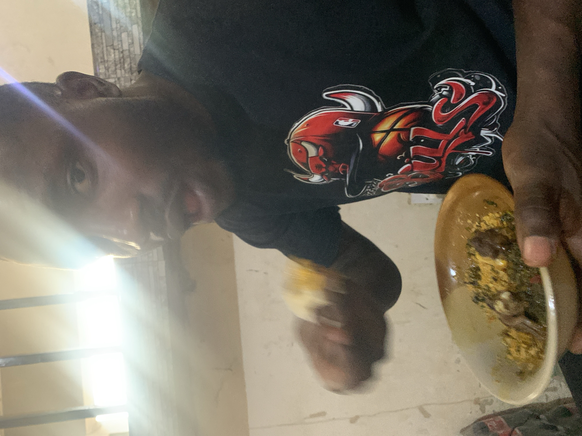 A person is holding a bowl of food and a portion of food in their other hand, seemingly about to eat. The person is wearing a black t-shirt with a red logo on it. The environment appears to be indoors. The location, according to the prompt, is Kabong, Nigeria.