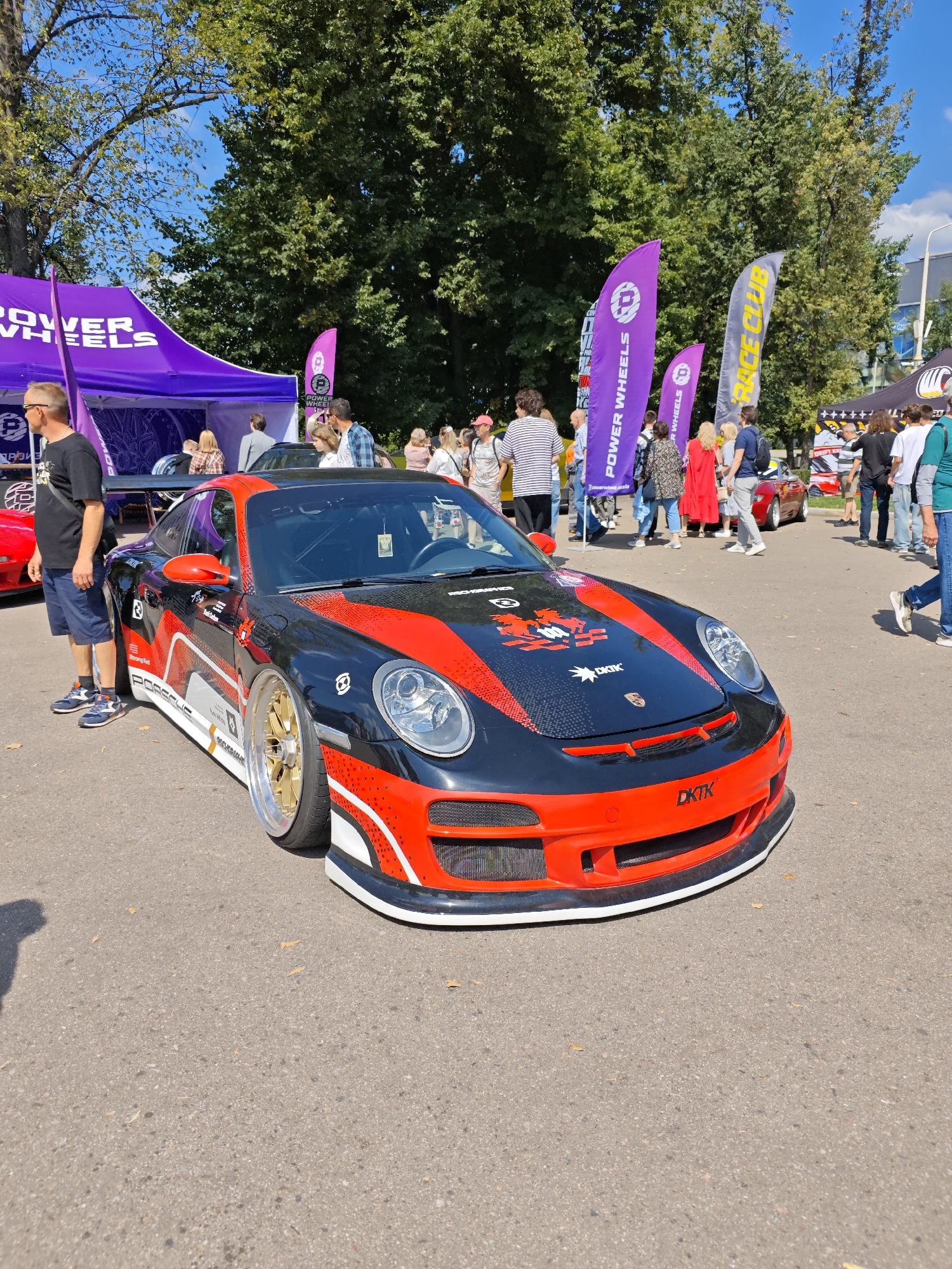 In Moscow, Russia, on a bright, sunny day, a black and red modified Porsche sports car is parked on an asphalt surface. The car has intricate racing decals and a prominent spoiler. A man wearing a black t-shirt and blue shorts stands to the left of the car, looking towards it. In the background, a crowd of people are gathered, some standing near tents and large flags. The flags display logos and text, including "POWER WHEELS" and "RACE CLUB." The environment appears to be an outdoor event, possibly a car show or exhibition, with trees providing shade in the distance.