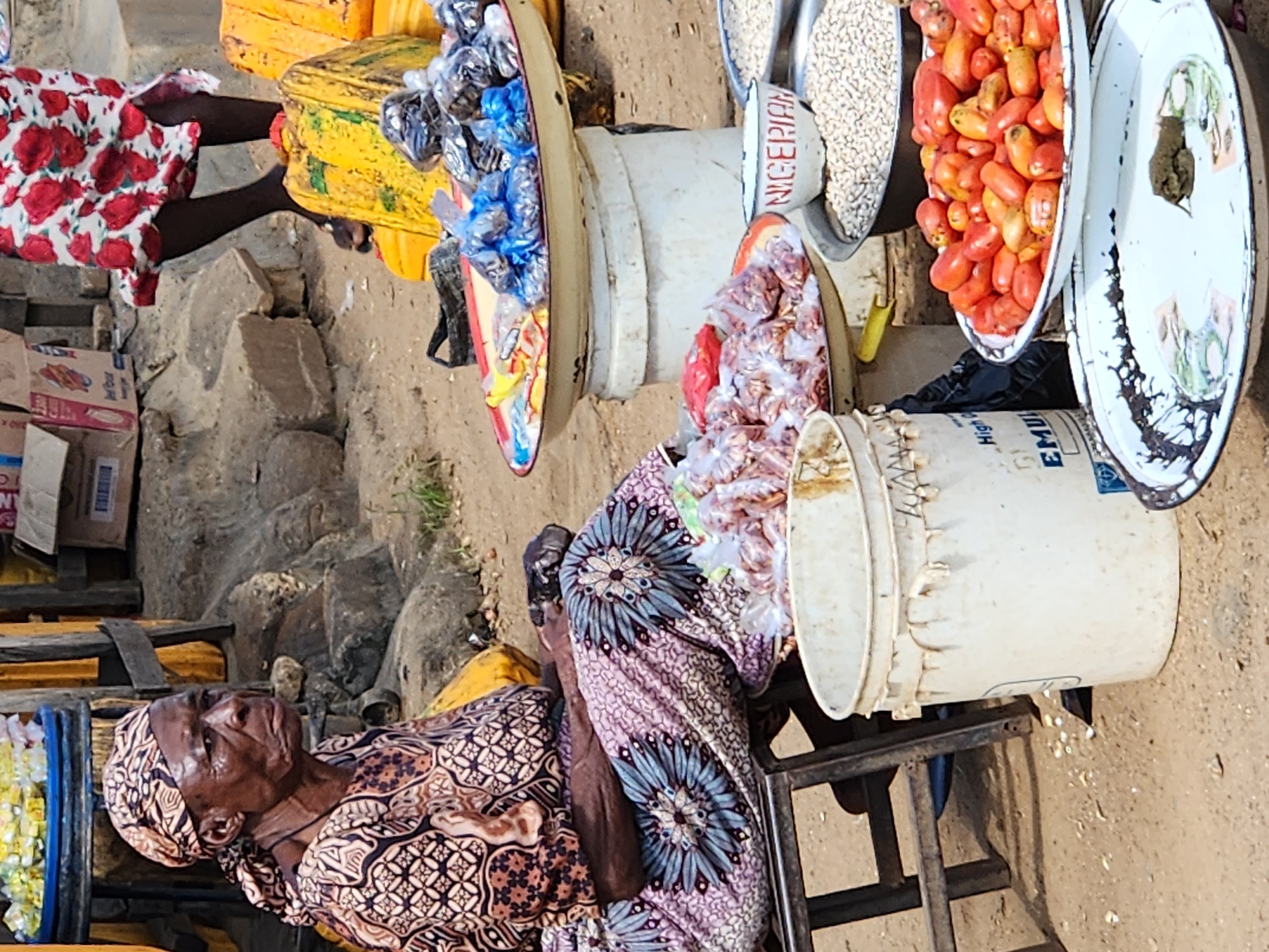 An elderly African woman wearing a patterned headwrap and a patterned dress sits on a stool at a market stall in Kabong, Nigeria. She is looking to her right. Her hands are resting on her lap. In front of her, on various trays and buckets, are various food items for sale. These include small red tomatoes, piles of dried beans, and plastic bags of what appear to be nuts or seeds. A white bucket with a label partially visible that reads "High Quality EMU" is to her left. The ground is sandy and dusty. In the background, other market stalls and people are visible. The overall scene depicts a bustling marketplace.
