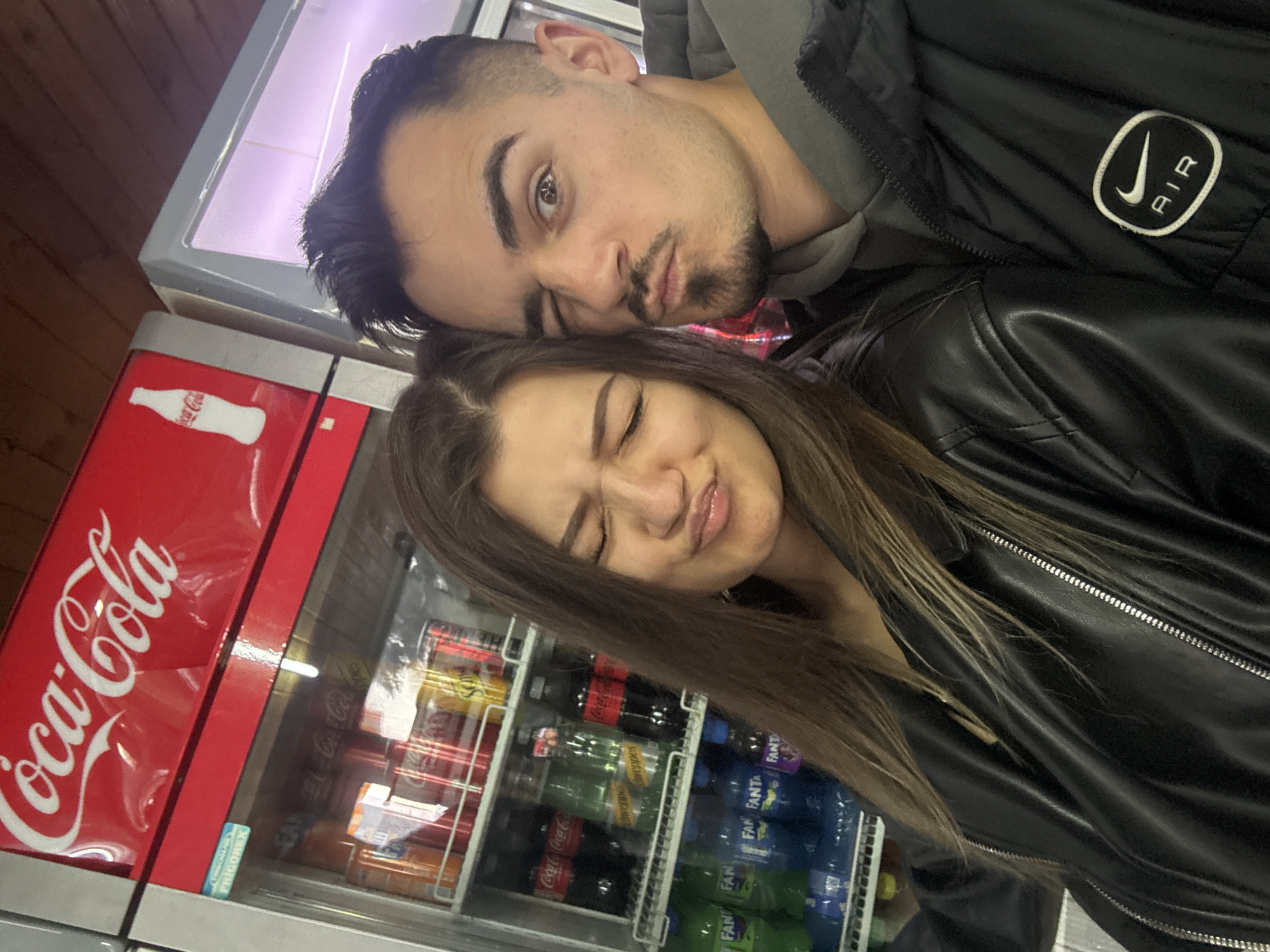 Two people, a man and a woman, are posing for a selfie in front of a Coca-Cola branded refrigerator. The man is leaning his head on the woman's, and both have their eyes closed and lips pursed, seemingly feigning a kiss. The man is wearing a dark jacket with a white "AIR" patch, and a gray hoodie can be seen underneath. The woman is wearing a black leather jacket. The refrigerator contains various beverages, including Coca-Cola, Fanta, and Schweppes. The OCR in the image also shows "FRIGOREX". The location is likely a convenience store or small market in Sofia, Bulgaria, based on the presence of the Coca-Cola refrigerator and common beverages.