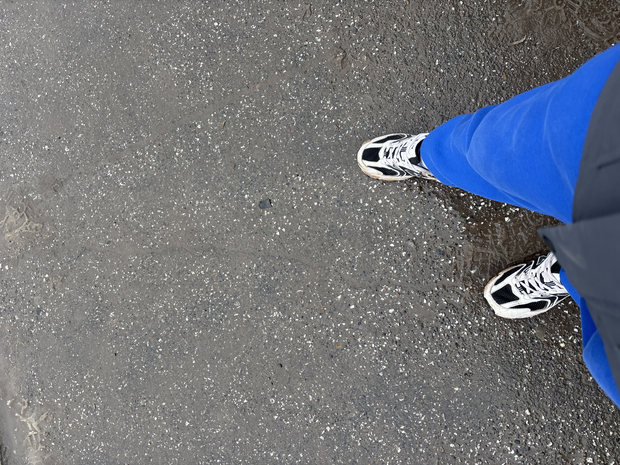 An eye-level shot depicts a person's feet on a wet asphalt surface. The person is wearing blue sweatpants and black and white running shoes. The asphalt is speckled with small white stones, indicating it is likely concrete. The lower right side of the image shows a portion of the person's gray jacket or coat. The exact location within Perm, Russia, cannot be determined from the image. The scene appears to be after a rain shower, evidenced by the wetness of the asphalt.