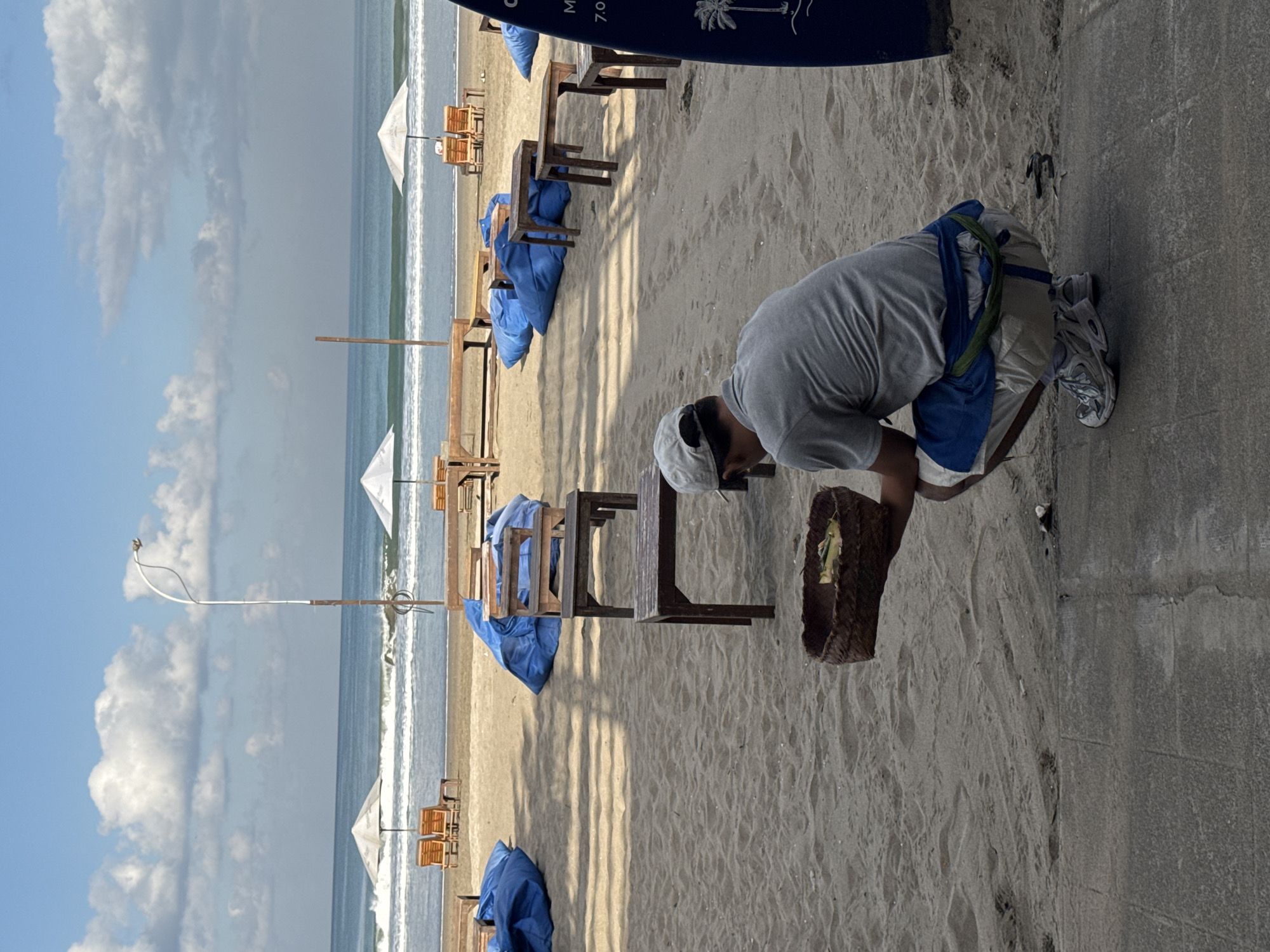 The image depicts a beach scene in Kuta, Indonesia, under a blue sky with scattered white clouds. The ocean with small breaking waves forms the horizon. On the sandy beach, multiple sets of wooden tables and chairs or loungers are arranged. Some seating areas are covered with blue tarpaulins or fabrics, while others have open white beach umbrellas. A tall white pole, possibly a lamppost, with curved wiring extends upwards from the sand on the left. Distinct shadows are cast onto the sand from an unseen overhead structure.
In the foreground, a man is squatting on a concrete path that borders the sand. He is seen from behind, wearing a grey short-sleeved t-shirt, light-colored shorts, a blue garment tied around his waist, and a white baseball cap worn backward. He has light-colored athletic shoes on his feet. The man is holding a brown, woven basket containing small, leafy items. To the right of the man, a blue surfboard or similar object stands upright, partially visible, featuring text including "7.0" and a palm tree graphic.