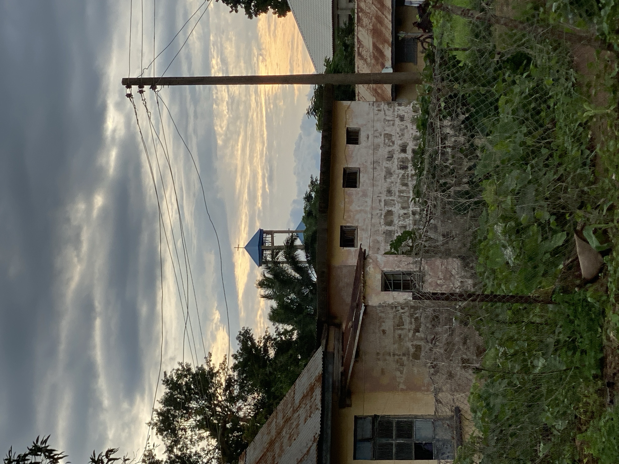 The image depicts an outdoor scene in Maigizo, Nigeria, at what appears to be dusk. The sky is filled with dramatic clouds, some dark and heavy, others illuminated by the setting sun with streaks of yellow and orange light. In the foreground, a wooden utility pole stands tall, supporting several electrical wires that stretch across the frame.
Behind the pole, the scene reveals a cluster of buildings. The closest structure has a weathered, light-colored facade with visible brickwork and a rusty corrugated metal roof. It has several small, square windows and one larger window with a gridded pane. To the left of this building, another structure with a white roof is partially visible. Further back, a taller building, possibly a tower, stands out with a blue, pointed roof topped by a cross. It is surrounded by lush green trees, including palm trees, which contrast with the man-made structures.
A chain-link fence, partially overgrown with green ivy and other foliage, runs along the right side of the image, separating the viewer from the buildings and vegetation. The overall impression is one of a somewhat aged or developing settlement, with a mix of utility infrastructure, residential or institutional buildings, and abundant natural growth. The time of day is suggested by the lighting in the sky, casting a warm glow through the clouds. There is no visible text in the image.
