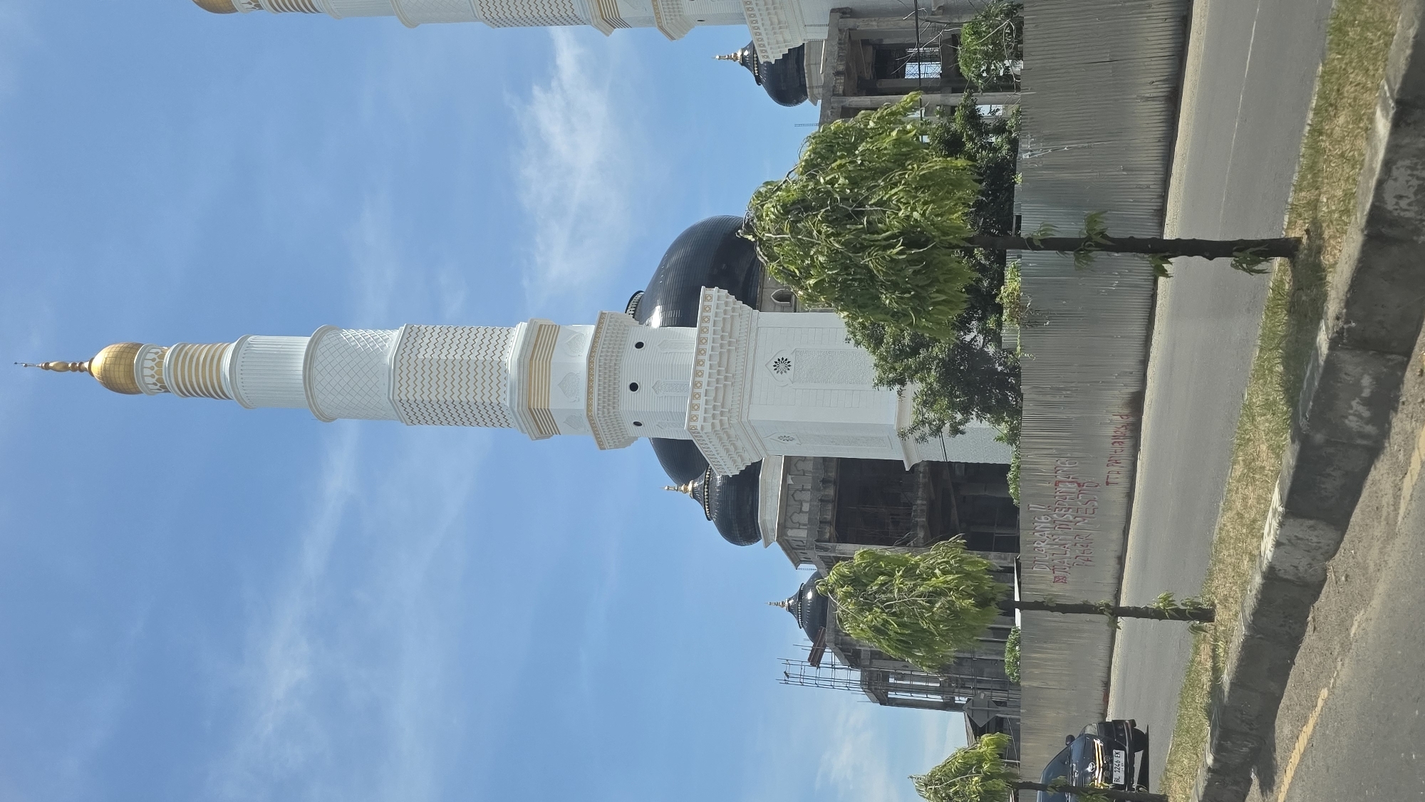 Here is a description of the image:
The image shows the Masjid Agung Islamic Center Lhokseumawe mosque under construction in Pantegajah, Indonesia. The mosque features a tall, white minaret with a golden spire, multiple black domes, and scaffolding. The foreground includes trees, a road, a corrugated metal fence, and a dark-colored car with license plate "BL 1249 EW." The sky is blue with scattered clouds. Graffiti is visible on the fence.