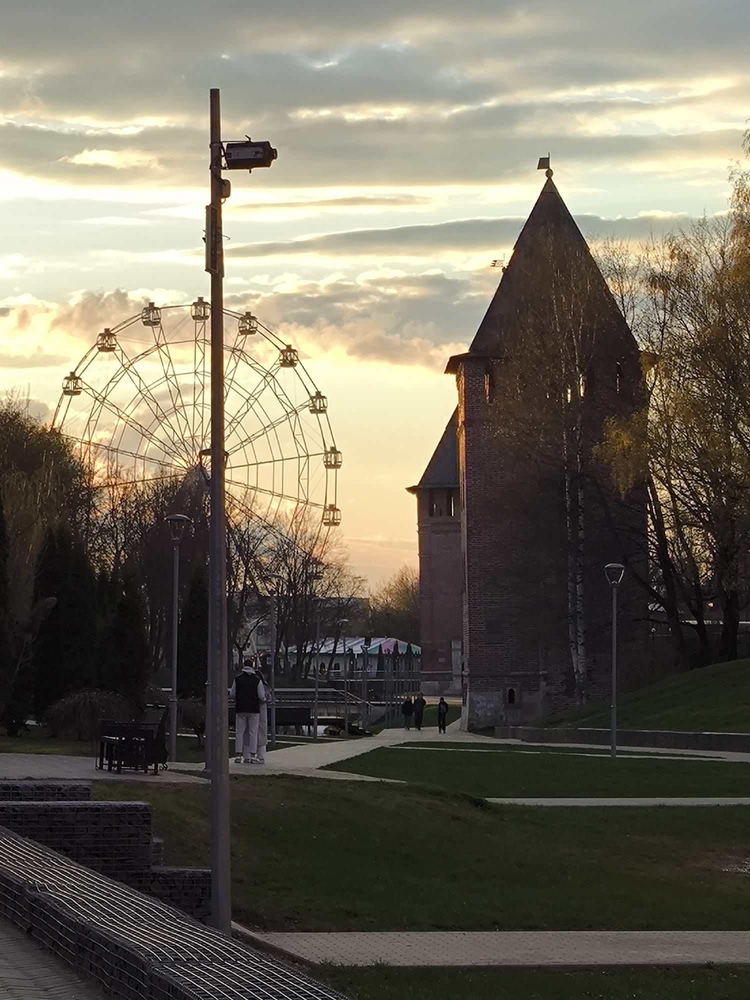 The image depicts a park scene with a Ferris wheel on the left and a brick tower on the right, set against a cloudy sky at sunset. A light pole stands in the foreground, slightly to the left. A person is walking along a path. The background includes trees and a structure with a colorful tent-like roof. The scene is in the city of Unknown, Unknown.