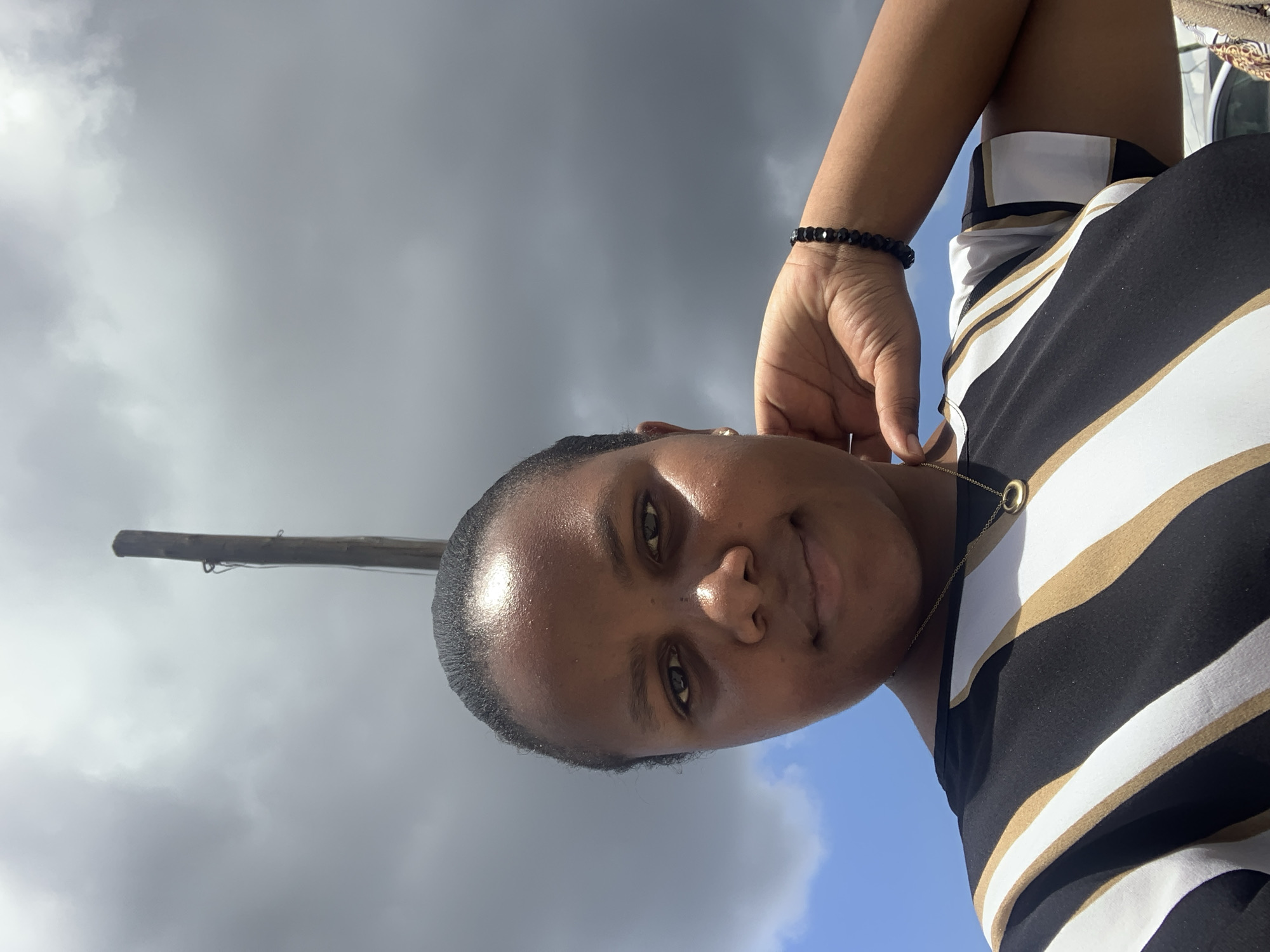 A woman with dark skin and hair pulled back is centered in a portrait-style shot. She wears a striped top in black, white, and gold, with a beaded bracelet on her left wrist and a gold necklace. Her left hand is raised near her face. The background is a sky with gray and blue clouds. A wooden pole is visible in the background, suggesting a possible urban setting, but without further context, definitive location within Dar es-Salaam, Tanzania, cannot be determined.