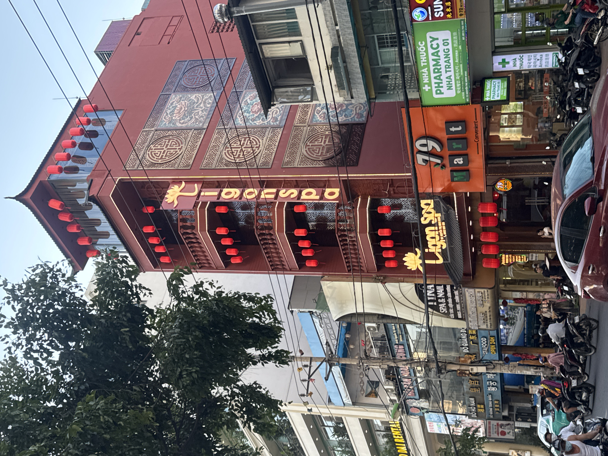 This is a street-level photograph taken in Nha Trang Ward, Vietnam, capturing a bustling commercial scene. The primary focus is a tall, multi-story building with a distinct red facade, decorated with traditional East Asian motifs and adorned with numerous red lanterns. The building houses businesses including "Ligon Spa" and "79 Mart." Adjacent to it, other buildings house various shops and pharmacies, indicated by signs such as "DẠI RENTAL," "SPA & NAIL," and "PHARMACY NHATRANG 01." The street is populated with people, many of whom are riding motorcycles, suggesting a busy urban environment. A few cars are also visible, including a red sedan in the foreground. Overhead, power lines crisscross the sky, a common feature in urban Vietnam. The time of day appears to be late afternoon or early evening, with the lighting suggesting a period shortly before sunset. The overall atmosphere is lively and indicative of daily commerce.