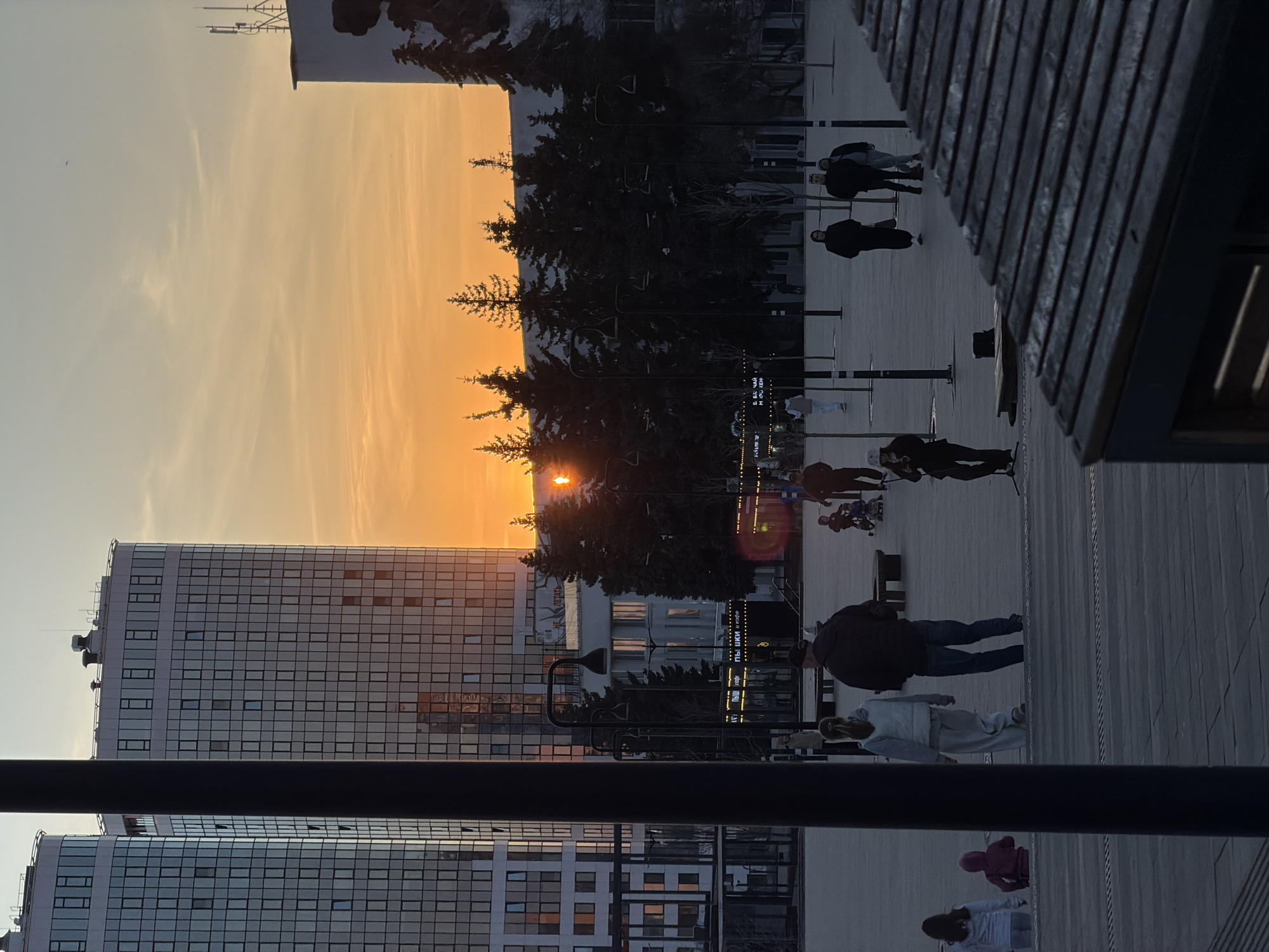 The image depicts a sunset scene in an urban environment. Tall buildings with numerous windows are visible to the left. In the center, the sun is setting behind a line of trees, creating a silhouette effect. People are walking and gathered in a public square with wooden decking. Streetlights are spaced along the square, and there is a partially obscured structure on the right side of the image.