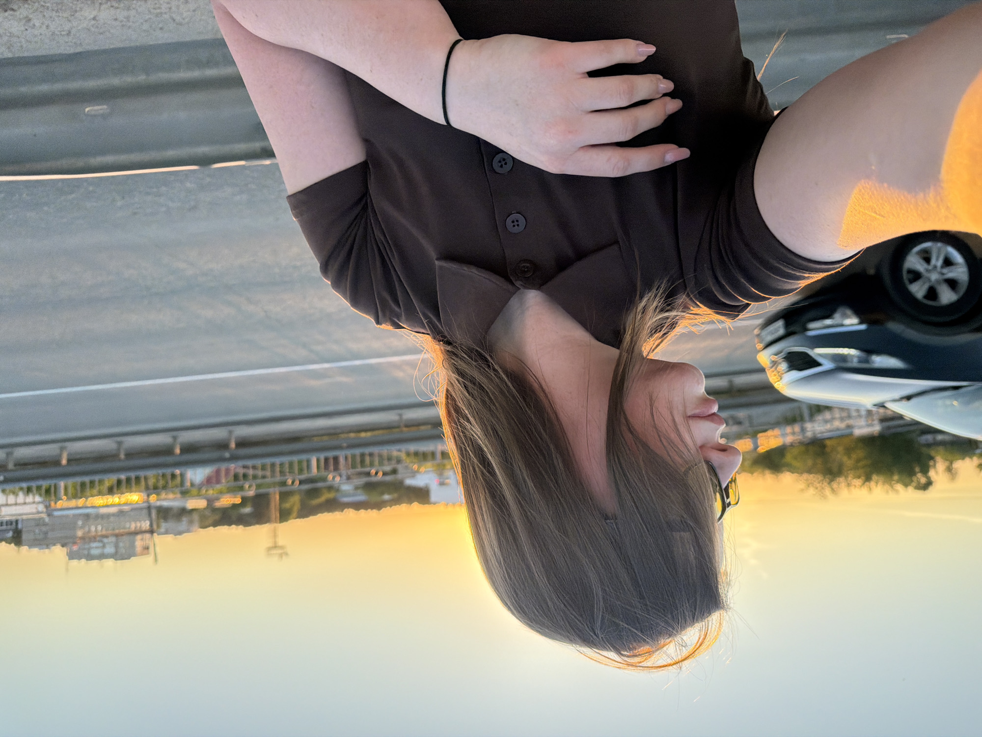A woman with long brown hair stands near a car at sunset in Chaikovsky, Russia. She is wearing a brown button-up shirt and glasses. The background shows a bridge, buildings, and a sunset sky.