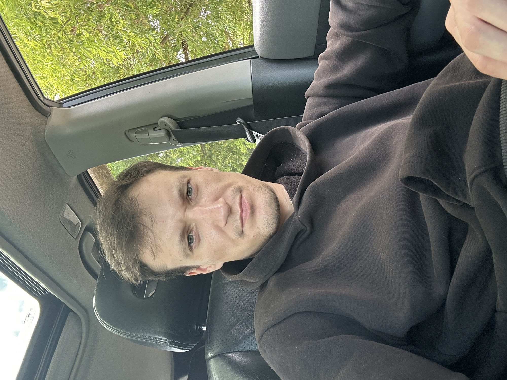 A man with short brown hair, light skin, and blue eyes is sitting in the driver's seat of a car. He is wearing a dark gray hooded sweatshirt. He is looking directly at the camera. The car interior includes a black leather seat, a gray dashboard, and a sunroof. Outside the car, green trees are visible. The location is Makiv, Ukraine.