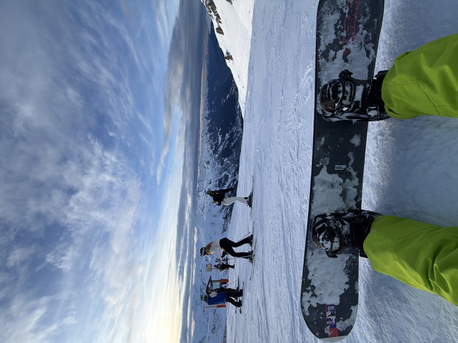 A snowboarder's point of view on a snowy mountain, with green pants visible. The snowboard is dark gray, covered in snow. Other skiers stand on the slope further down the hill. Mountains stretch out into the distance under a cloudy sky, creating a panoramic winter scene.