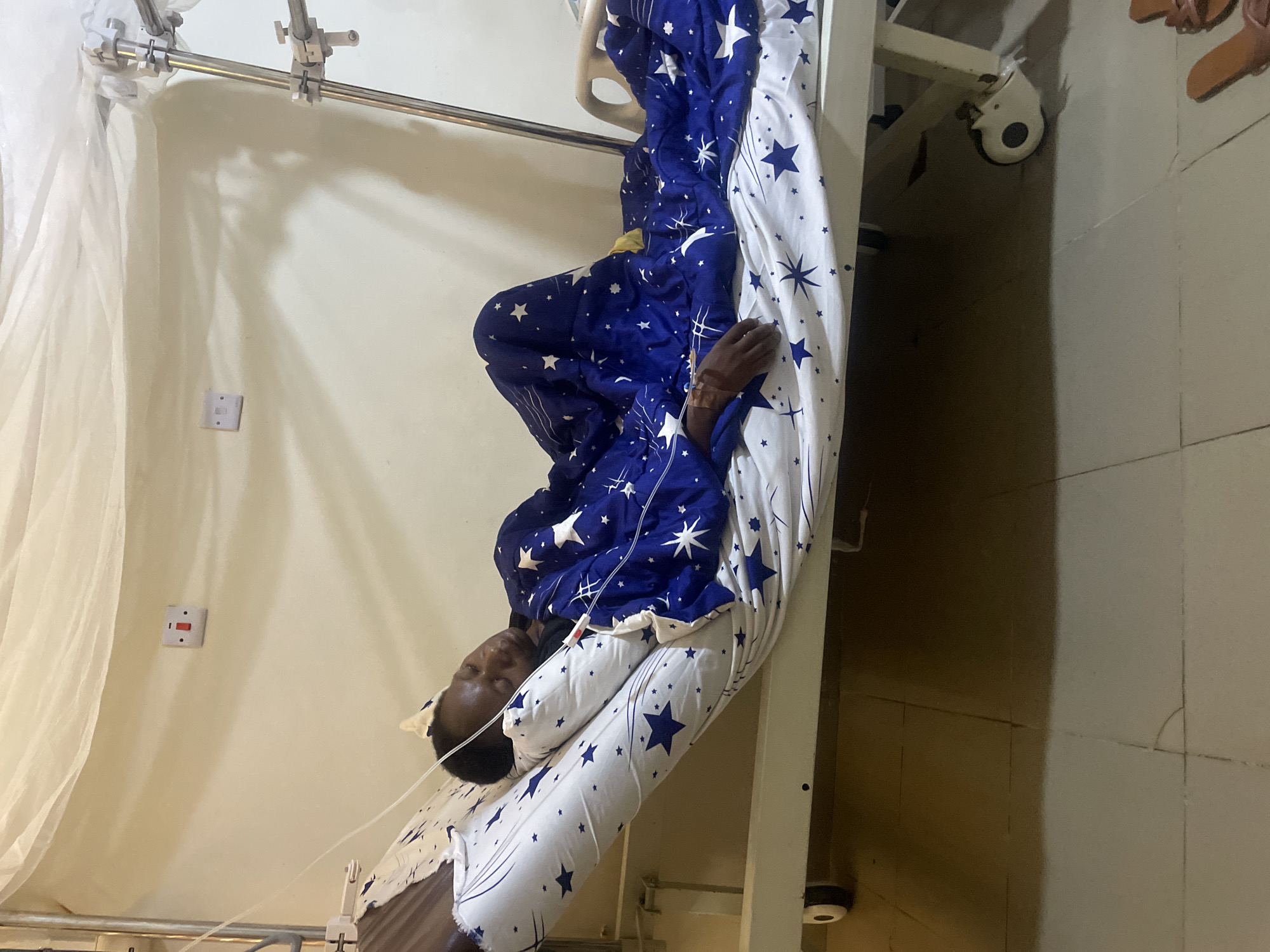 A Black person lies in a hospital bed in what appears to be a medical ward. The person is covered with a blue and white star-patterned bedspread and has an IV drip attached to their arm. The setting includes standard hospital equipment, such as adjustable bed rails and IV stand. The hospital room has tiled floors and walls, with visible electrical outlets. There is no explicit evidence in the image to corroborate the precise location as Gwong, Nigeria.
