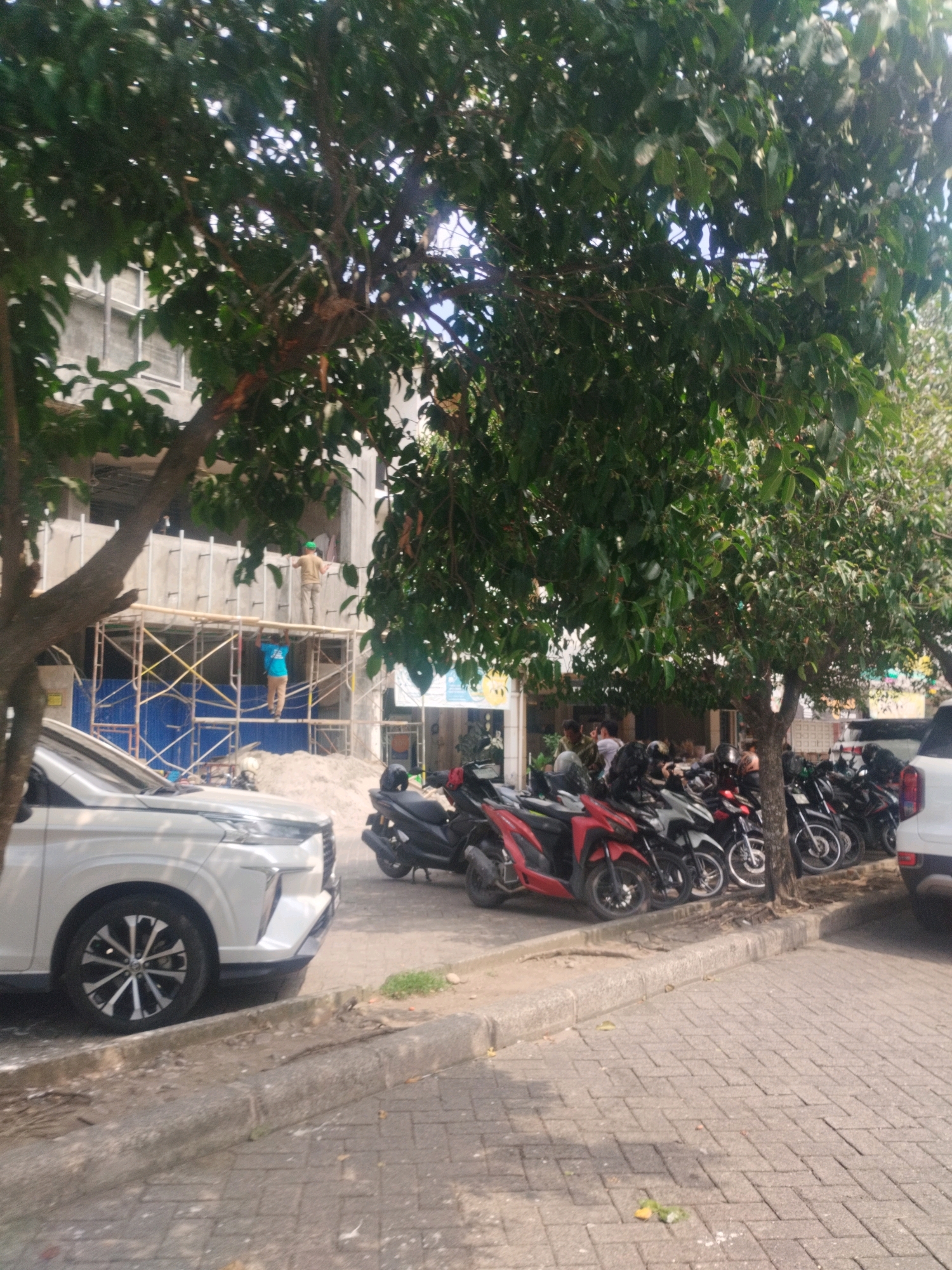 This image captures a street scene in Percut Sei Tuan, Indonesia, with a construction site and parked motorcycles. In the background, a multi-story building is under construction, with scaffolding erected around the upper floors. Two construction workers, one in a yellow shirt and green helmet, and another in a blue shirt, are visible on the scaffolding, seemingly engaged in work.
In the foreground, a white car is parked on the left side of the frame. To the right of the car, a row of motorcycles is parked along the sidewalk. These motorcycles are of various colors, including red and black, and many are covered with dark-colored seat covers. A large tree with lush green leaves partially obscures the view of the construction site and some of the parked motorcycles. The ground is paved with interlocking bricks, and a concrete curb separates the road from the sidewalk. The overall impression is a bustling urban environment with ongoing development and typical street activity.