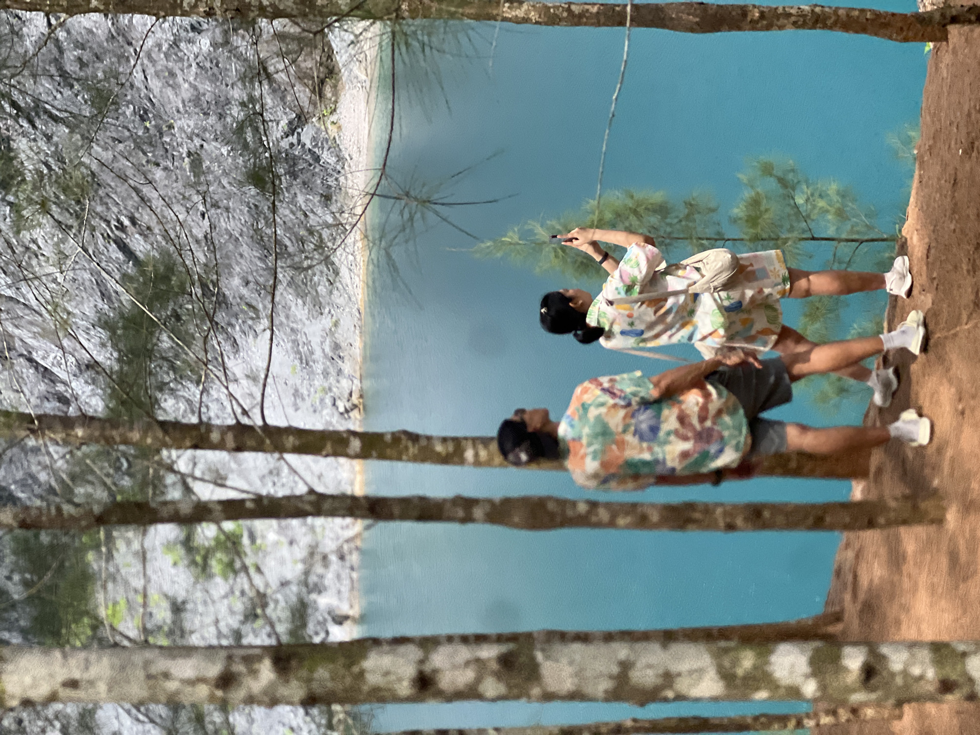 Here is a description of the image:
The image depicts two people standing on a reddish-brown, possibly earthen, ledge, overlooking a body of bright blue water. The location is Ban Nai Wang, Thailand. The body of water is likely a lake, with a white, rocky or sandy shoreline visible on the opposite side. Several tall, thin trees with light-colored trunks and sparse branches frame the scene in the foreground.
The first person, presumed to be a woman, is wearing a light-colored patterned shirt, light shorts, white shoes, and white socks. She is also carrying a small bag across her body. She is holding a phone in the air.
The second person, presumably a man, stands slightly behind her. He is wearing a patterned shirt, dark shorts, white socks, and white shoes.
The sky and sunlight are reflected by the blue water. The environment looks natural.
