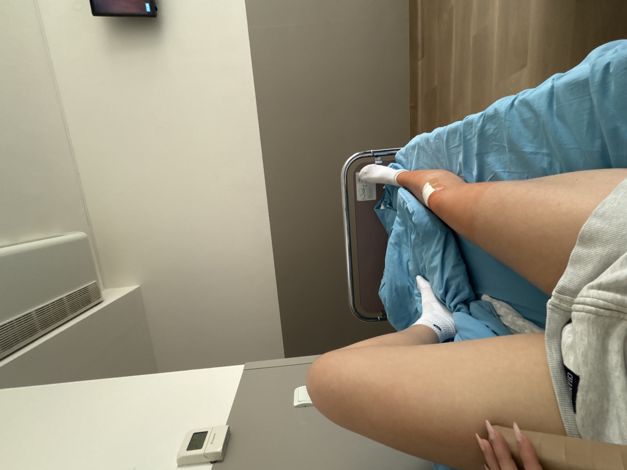 A first-person view of a person lying in a hospital bed. The individual has white socks on and a bandage on their leg. The bed has blue sheets. A bedside table is visible with a control panel on it. In the background, there's a wall with an air conditioning unit and a television mounted high up. The scene is likely taking place in a hospital room in Sofia, Bulgaria, based on the available information.