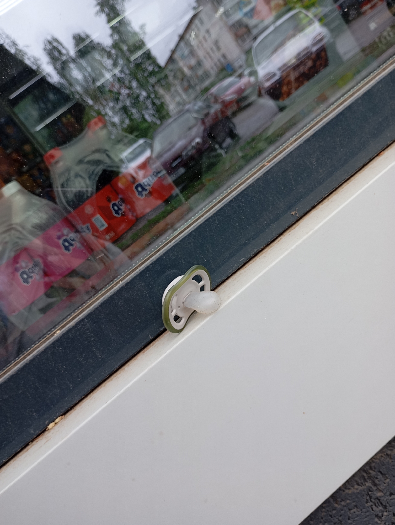 Observed outdoors in an urban environment of Unknown, Unknown, this eye-level shot showcases a window reflecting the exterior. Within the window frame, packages of "Aquolina" brand products are visible. Affixed to the window is a white and green pacifier. The window frame is white and gray. The reflection shows cars and greenery. The surface adjacent to the window is white.