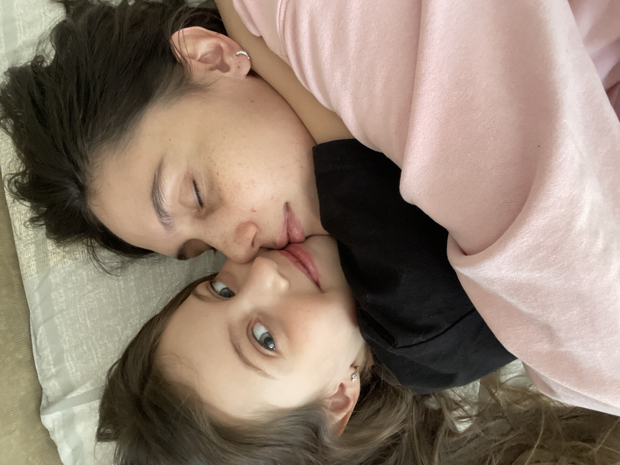 This image depicts two individuals, an adult female and a young female, in a close embrace, located in the city of Izhevsk, Russia. The adult female, positioned on the right, has dark hair, closed eyes, and visible freckling on her cheek. She wears a light pink long-sleeved garment and a silver-colored hoop earring in her right ear. Her face is pressed against the left side of the young female's head and cheek. The young female, positioned on the left, has light brown hair and blue-green eyes open and looking towards the camera. She exhibits a faint smile. She wears a black garment and a gold-colored, flower-shaped earring in her left ear. Her left arm is wrapped around the adult female's upper body. Both individuals are lying down on what appears to be a light-colored, patterned bed or couch linen. The interaction suggests a moment of close physical affection.