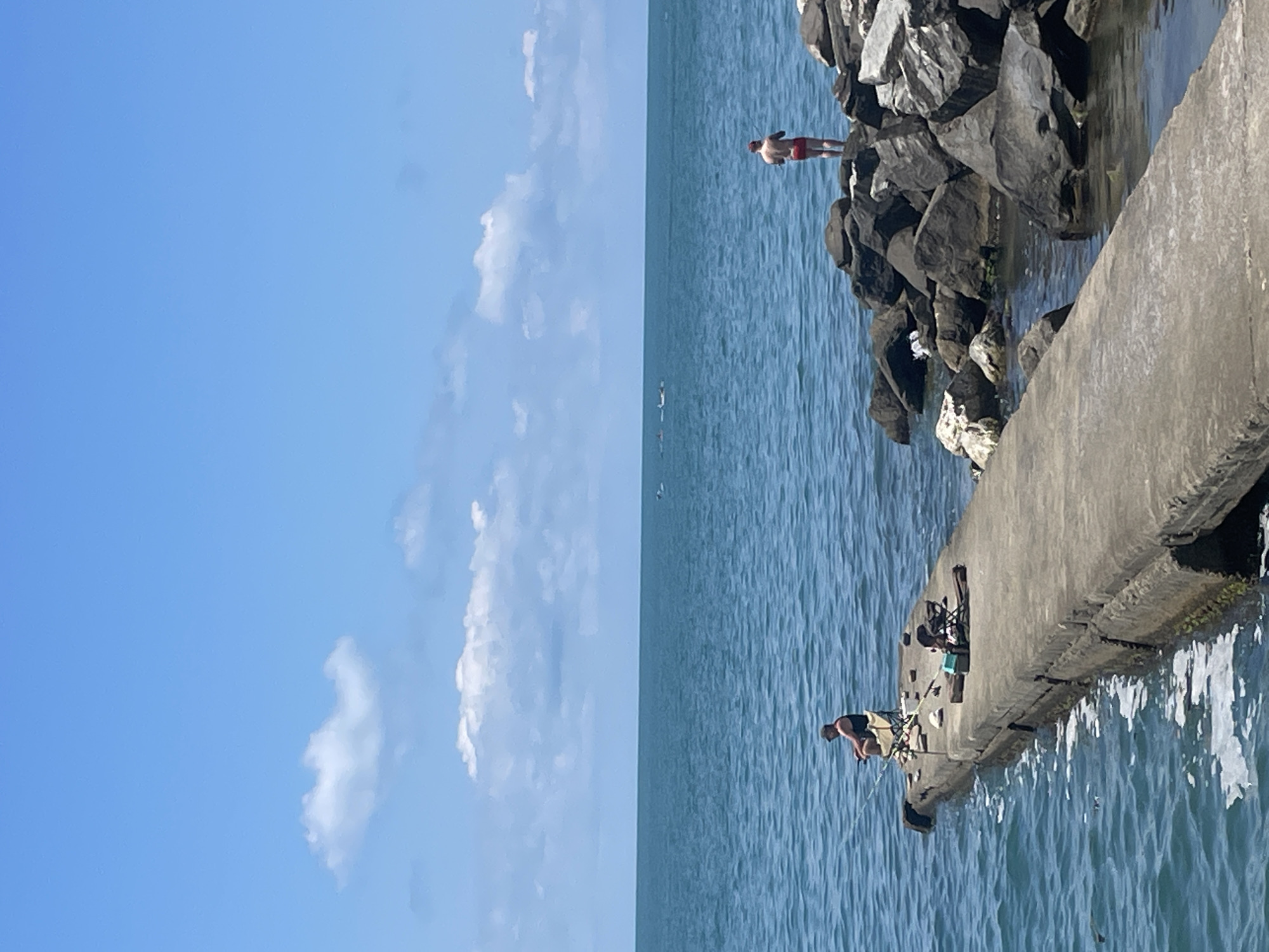 The image depicts a scenic view of the Black Sea near Sochi, Russia, on a clear, sunny day. The vast expanse of the blue sky with scattered white clouds dominates the left side of the frame. The right side of the image transitions to the deep blue, rippling water of the sea, bordered by a rocky breakwater and a concrete pier.
On the rocky breakwater, a person is visible standing near the edge, wearing red shorts. On the concrete pier, another person is engaged in fishing, with fishing rods and a tackle box set up. The pier appears weathered, with visible cracks and erosion. The water is calm, with gentle waves lapping at the pier and rocks.
The scene suggests a leisurely day by the sea, with activities like sunbathing and fishing taking place. The bright sunlight indicates it's likely midday. The location cues, such as the rocky coastline and the style of the breakwater, are consistent with coastal areas. No visible text is present in the image. The overall mood is peaceful and summery.