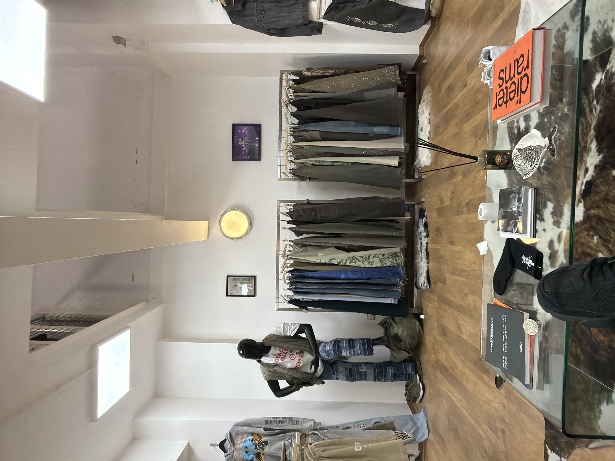 This is a photograph of the interior of a clothing boutique. The walls and ceiling are white, and the floor is wooden. Two clothing racks display a variety of apparel, including jeans and other garments. A mannequin is dressed in clothing on display.
On a glass-topped table near the clothing racks are several items, including books, a small decorative dish, a diffuser, and sneakers. An orange book titled "dieter rams" is prominently featured.
The image lacks specific indicators to confirm it is in London, United Kingdom. More visual clues related to the location would be needed for confirmation.