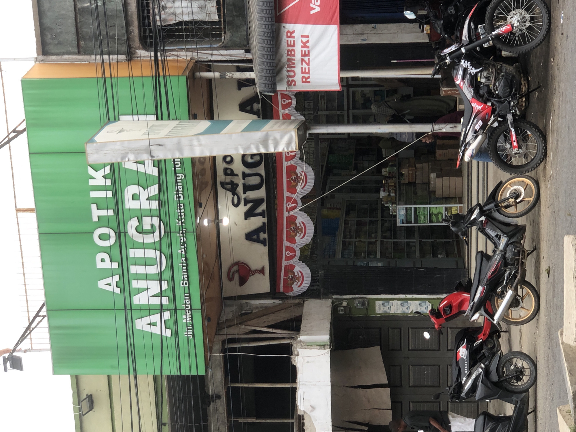 The image depicts the exterior of an "APOTIK ANUGRAH" pharmacy located in Titibaro, Indonesia. The pharmacy features a large green sign with white lettering that reads "APOTIK ANUGRAH". Below the main sign, smaller text indicates the address: "Jln. Medan Banda Aceh, Kuta Blang, idi".
In front of the pharmacy, several motorcycles are parked on the street. Two black scooters are on the left, with a red scooter parked behind them. A third motorcycle, a red and black dirt bike with gold rims, is positioned in the foreground on the right.
The entrance to the pharmacy is visible, adorned with red and white banners featuring an emblem. Inside, shelves stocked with products are discernible through the open doorway. A person is partially visible entering the pharmacy, and another person in a green headscarf is standing inside. On the right side of the image, a red and white banner with the text "SUMBER REZEKI" is displayed. The overall environment suggests a street scene in a commercial area.