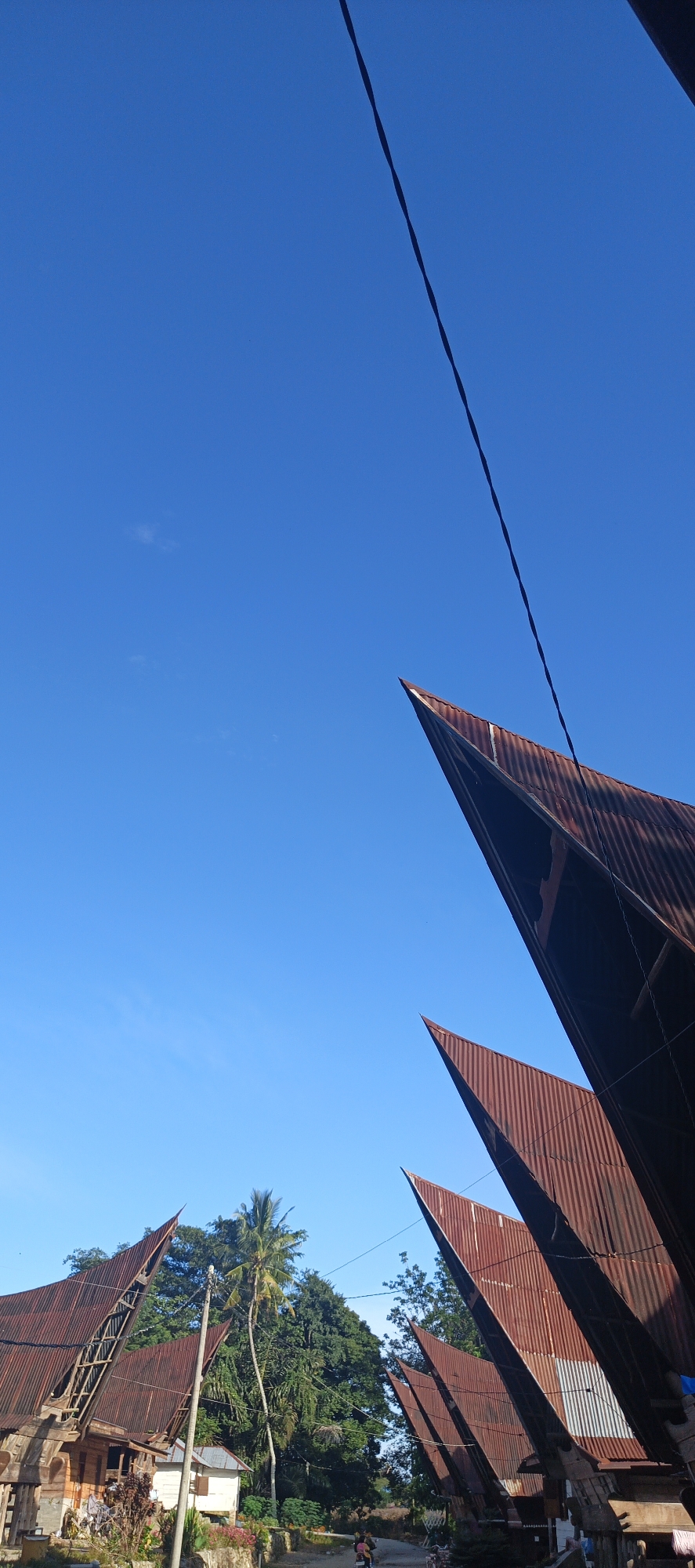 The image captures a scene in Cinta Dame, Indonesia, characterized by traditional houses with distinctive peaked, rusty metal roofs. The sky is a clear, vibrant blue, with a single, thin power line stretching diagonally across the upper portion of the frame. In the foreground, several of these unique roofs are prominently visible, their corrugated texture and reddish-brown hue contrasting with the bright sky.
To the left of the frame, one of these houses is more fully visible, showing wooden construction and a porch area where a few people are seated. A smaller white building stands nearby. Behind the houses, lush green trees and palm trees form a dense backdrop. A narrow, unpaved road runs through the scene, with a person on a motorcycle visible in the lower center of the image, moving away from the viewer. The overall impression is one of a rural or semi-rural setting, with architectural elements unique to the region.