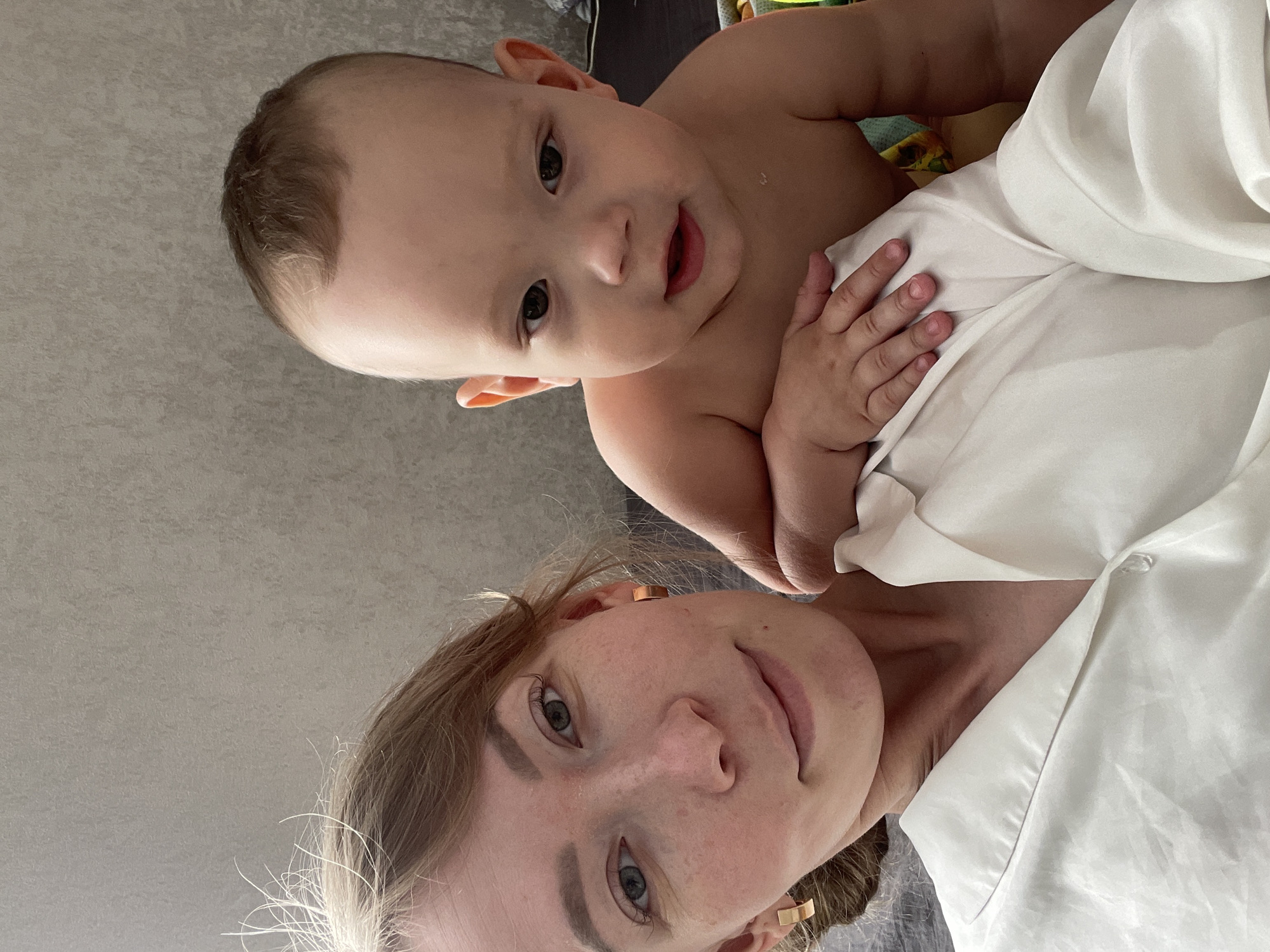 A close-up, slightly rotated image features a fair-skinned woman and a baby. The woman has light brown hair, fair skin with visible freckles, and is wearing what appears to be a white shirt. She looks directly into the camera. The baby, positioned above her, also has fair skin and brown hair. The background consists of a neutral-toned wall or surface. While the city of Perm, Russia, is mentioned, there's no discernible visual information within the image to definitively confirm the location.