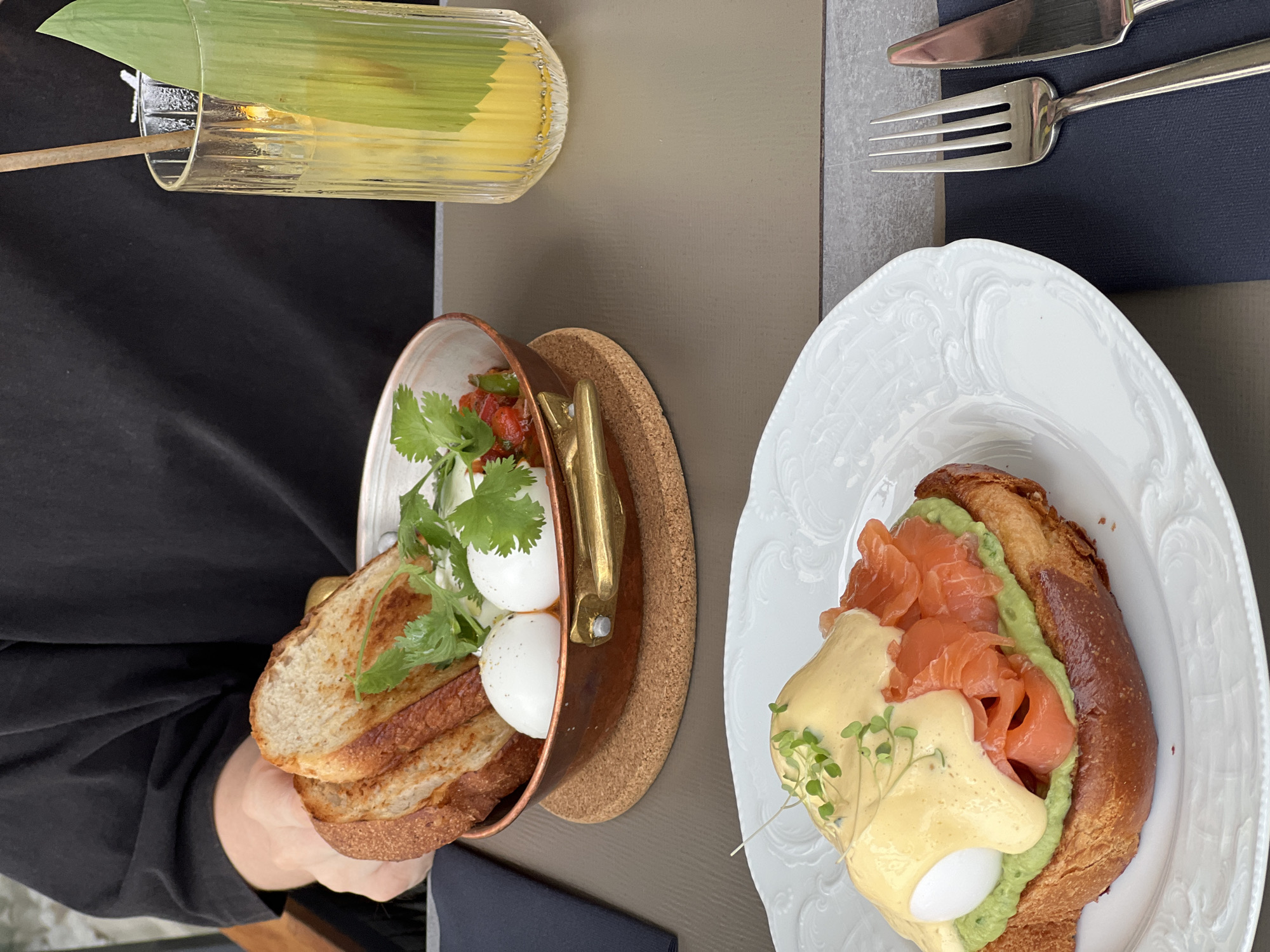 The image shows a table setting at a restaurant, likely in the city of Boreti, Montenegro, based on the potential context of the scene. A person is holding a copper pot containing slices of toasted bread, poached eggs, a tomato-based relish, and sprigs of cilantro. Adjacent to the pot is a white, ornate plate holding a croissant topped with avocado, smoked salmon, Hollandaise sauce, and microgreens. A fork and knife are placed to the right of the plate on a black napkin. To the left of the copper pot, there is a tall, narrow glass containing a light-colored liquid, possibly a cocktail or juice, garnished with a leaf. The tabletop has two distinct sections: a light grey, stone-like surface and a light brown, solid surface. The person holding the pot is wearing a black shirt. The food appears to be brunch or a similar meal.