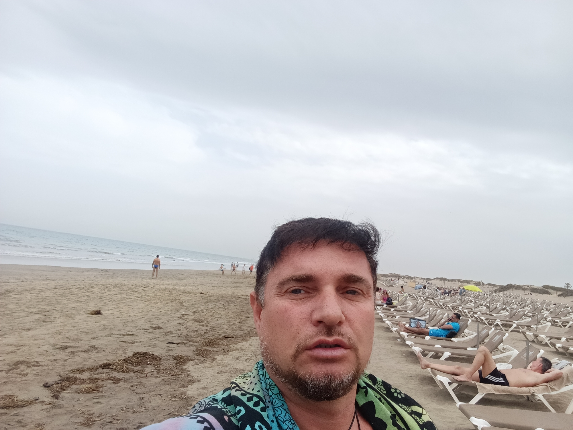 The image is a selfie taken on the beach in Maspalomas, San Bartolomé de Tirajana, Spain. The camera is held by a man with short dark hair, a salt-and-pepper beard, and a patterned shirt. Behind him is a sandy beach with several people and rows of sun loungers. In the background, people are walking along the shoreline. To the right, a large number of sun loungers are arranged in rows, some occupied by people sunbathing. The sky is overcast. The sea is calm and visible in the distance.