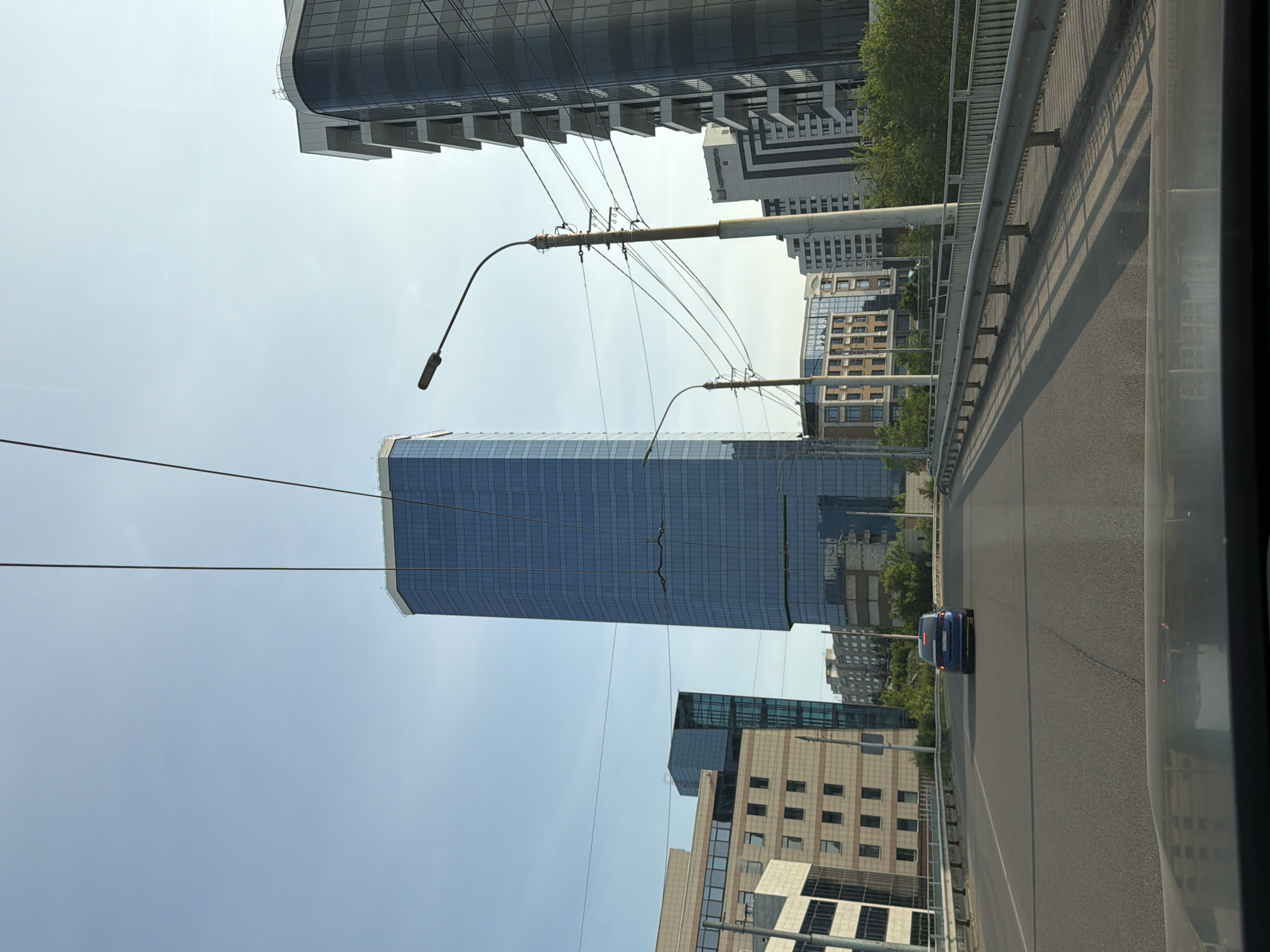 The image shows a view from a vehicle traveling on a road, likely a bridge, with modern skyscrapers in the background. The scene is dominated by tall buildings with glass facades, characteristic of a city environment. A blue car is visible on the road ahead, moving in the same direction. Power lines and streetlights are also present, suggesting an urban infrastructure. The sky is clear and bright, indicating daytime with fair weather. The orientation of the image is such that the buildings appear to be stretching upwards from the ground, and the road runs horizontally across the frame. The overall impression is one of a contemporary cityscape, possibly during a commute or travel through a developed area. There is no visible text in the image.