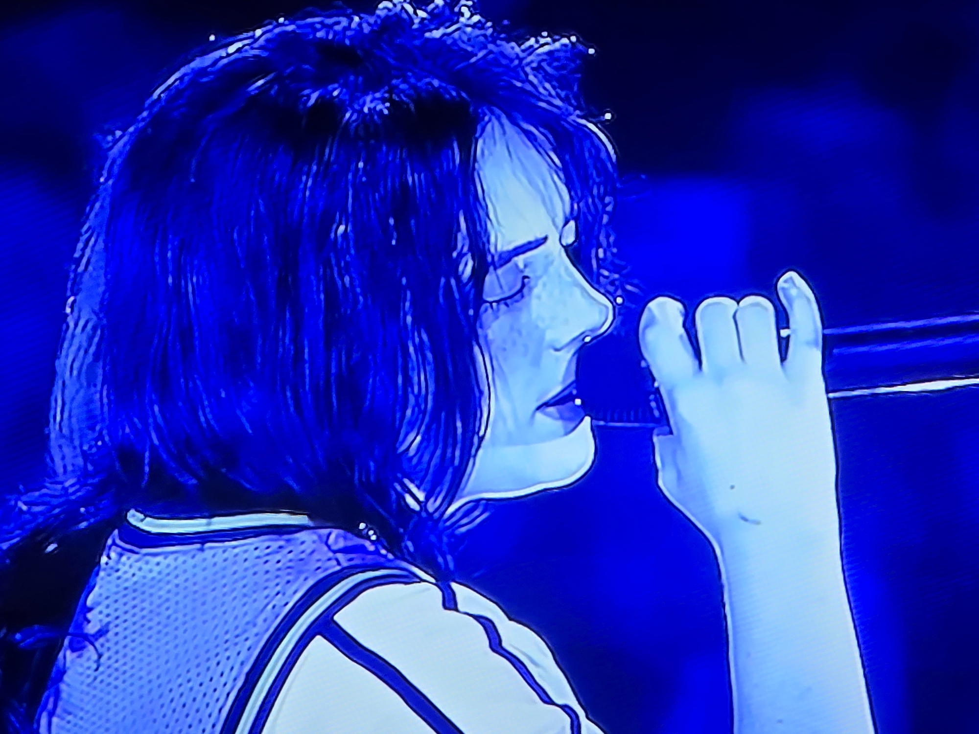 A close-up, side profile image of a young woman singing into a microphone under intense blue lighting. Her eyes are closed, and her face is slightly tilted upwards. She has dark, shaggy hair and is wearing a light-colored garment with dark trim, possibly a jersey. Her left hand is visible, holding the microphone, with some small marks on her skin. The background is a dark, blurry blue. The image quality is somewhat grainy, suggesting it may be a photograph of a screen or taken in low light conditions. There is no discernible location in the city of Satbayev, Kazakhstan within the image.