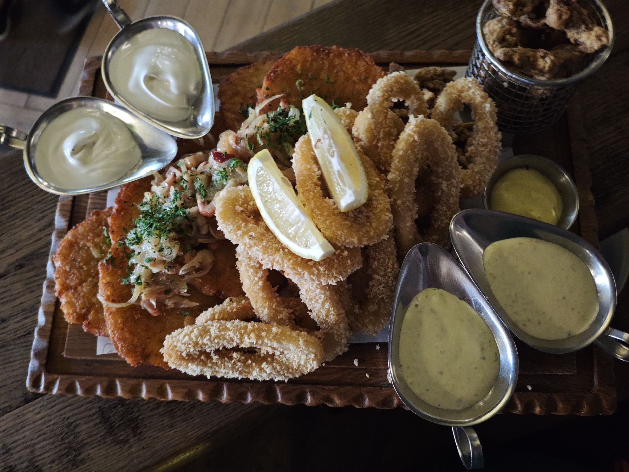 A platter of food, including fried calamari rings, potato pancakes, and what appears to be a small metal basket of fried items, is presented on a wooden board on a dark wooden table. Two metal gravy boats filled with a creamy yellow sauce are positioned in the foreground, and two smaller metal dishes containing white sauces are in the upper left. Two lemon wedges are placed on top of the calamari. The platter is also garnished with fresh parsley and what looks like shredded onions and herbs. The overall lighting is dim, suggesting an indoor setting, possibly a restaurant. No people are visible in the image. The context of Kyiv, Ukraine, is not visually inferable from the image itself.