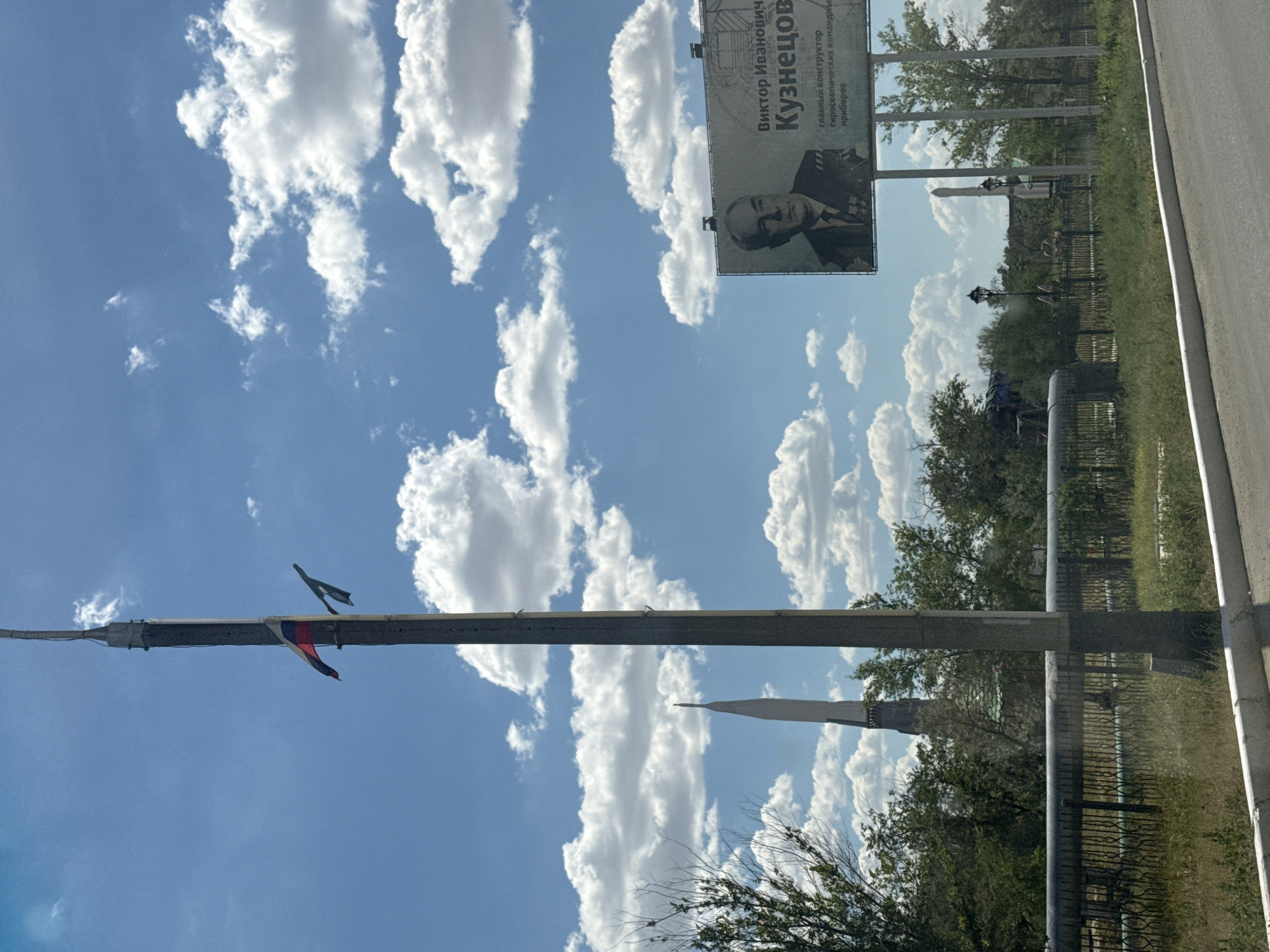 This is a photograph taken outdoors with a bright, cloudy sky dominating the upper portion of the frame. A tall, concrete lamppost runs diagonally across the center of the image. Two small flags, one appearing to be the Russian tricolor and another dark one, are attached to the lamppost. In the upper right, a billboard is visible, oriented vertically. It features a portrait of a man wearing a suit and medals, with Russian text above him. The text reads: "Виктор Иванович Кузнецов" (Viktor Ivanovich Kuznetsov) and below that, "главный конструктор гидроскопических командных приборов" (chief designer of gyroscopic command instruments). The billboard is likely advertising or commemorating this individual.
The lower portion of the image shows a green, grassy area with trees and a fence. In the background, a tall, slender structure with a pointed top, possibly a monument or a part of a larger building, is visible against the sky. The overall scene suggests an outdoor location, possibly a park or a public space, with a clear day and moderate cloud cover. The orientation of the image is rotated 90 degrees counter-clockwise. The context of Baikonur, Kazakhstan, suggests a setting related to space exploration or aerospace industry, given Kuznetsov's title as chief designer of gyroscopic command instruments. The time of day appears to be daytime, with good lighting.