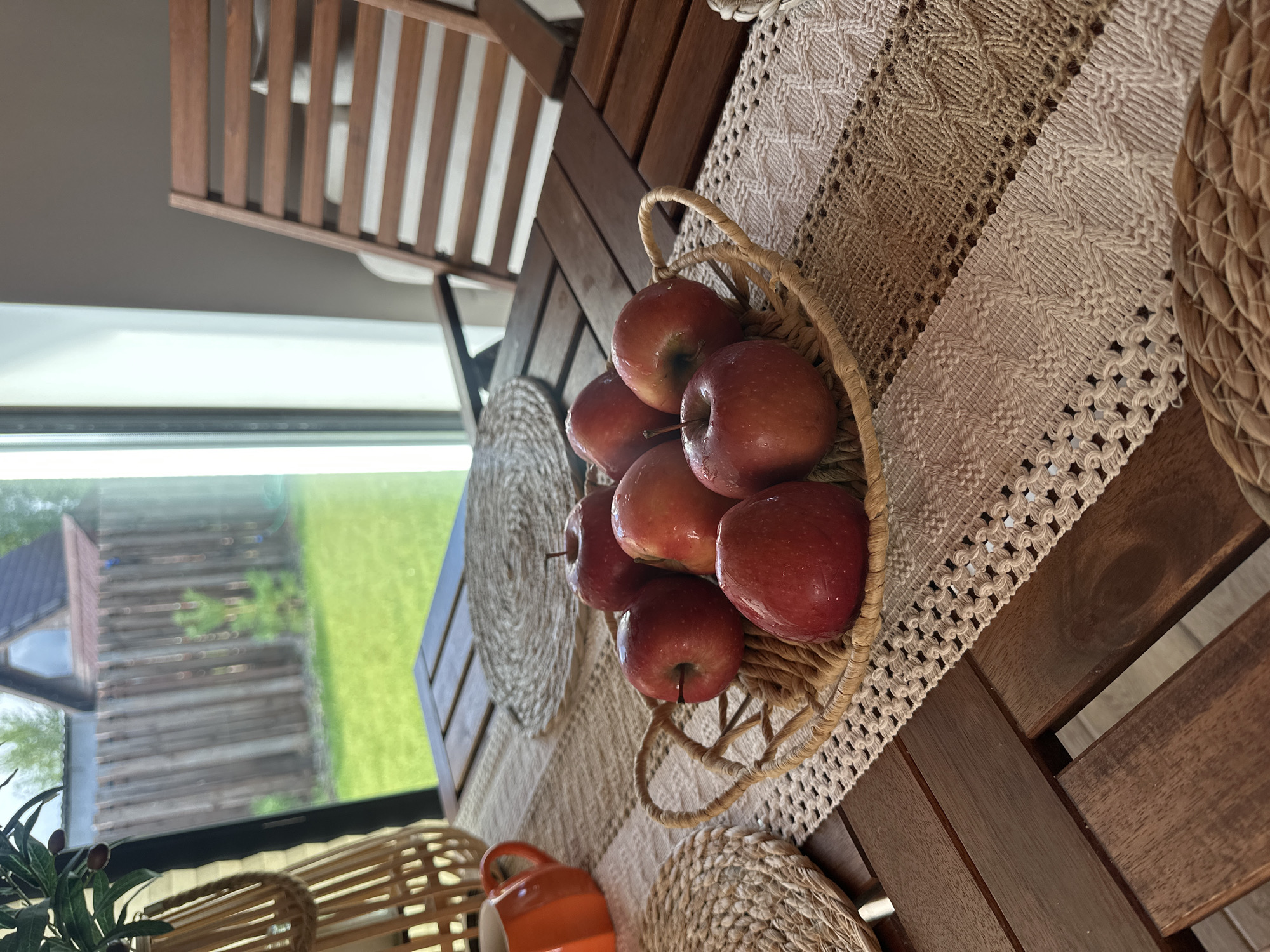 A close-up, eye-level shot focuses on a woven basket filled with seven red apples, resting on a table runner with a macrame and striped pattern. The basket and runner are on a dark wood slatted table. In the background, through a large window, a green lawn, wooden fence, and a portion of a house are visible. To the left, a portion of a woven decorative item and an orange ceramic pitcher are visible. To the right, the arm and seat of a wooden chair are present. The lighting is natural, coming from the window.