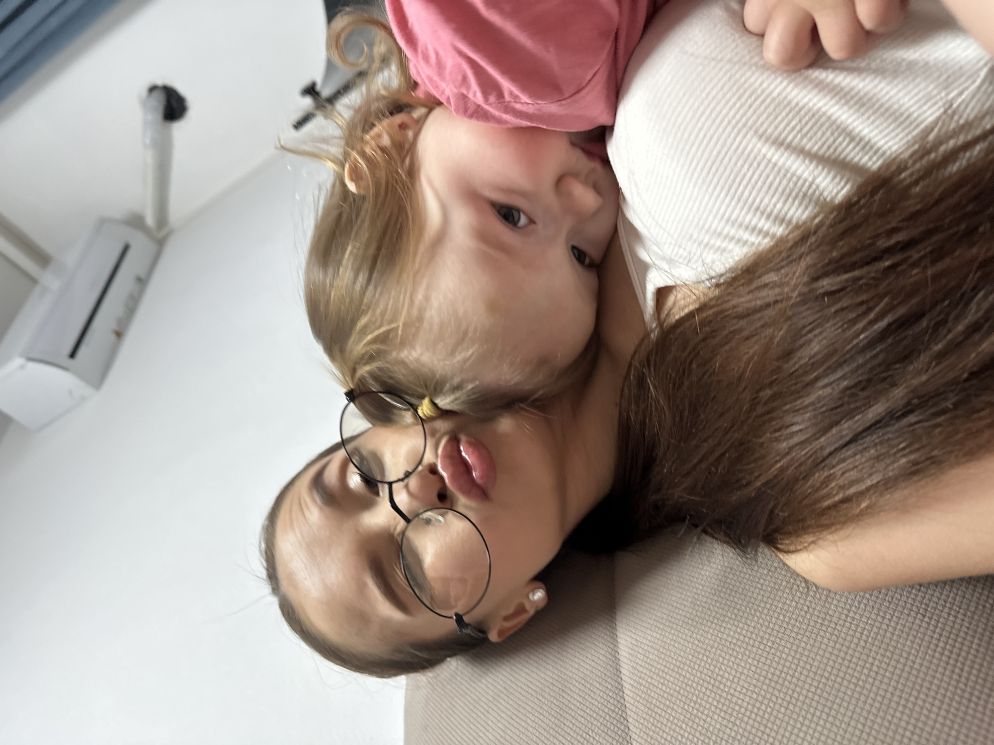 This sideways-oriented portrait captures a tender and playful moment between an adult and a young child in an indoor setting.
The adult, positioned in the lower right of the frame, has long dark brown hair, round-framed glasses, and is wearing a light-colored, possibly ribbed, top. She has a pearl earring in her right ear and is making a playful expression with pursed lips, looking towards the camera.
Resting closely against her, with their heads touching, is a young child, likely a toddler or pre-schooler. The child has light brown or blonde hair and is wearing a pink top. With wide, curious eyes, the child is looking directly at the camera, conveying a sense of innocence and engagement. One of the child's hands is visible resting on the adult's white top.
The subjects appear to be lying on a textured, light brown or grey couch or sofa, which occupies the lower right portion of the image. The background features a simple white wall with an air conditioning unit mounted high up. A vertical pipe or conduit extends from the ceiling near the AC unit.
The image conveys a warm, affectionate, and intimate family moment. The unique angle adds a casual and personal feel to the photograph. No specific time of day or outdoor weather can be determined from the indoor setting. There is no visible text within the image. While the location is contextualized as Yekaterinburg, Russia, no specific visual cues in the photo itself directly indicate this city.