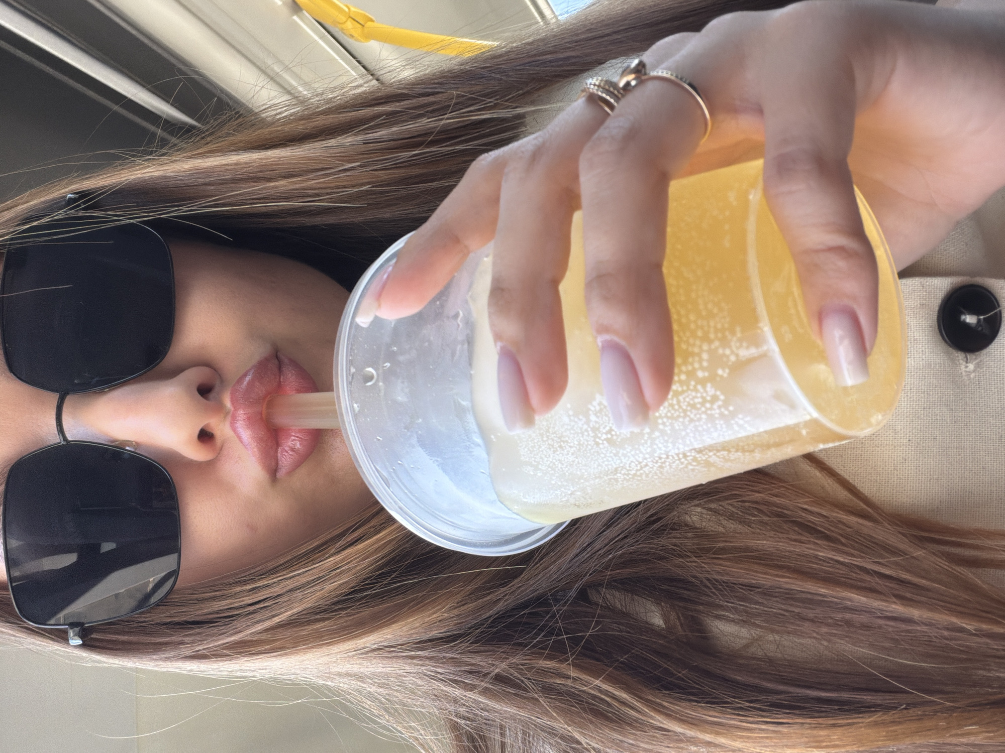 A woman with long brown hair and sunglasses is drinking from a clear plastic cup filled with a yellow beverage and a straw. She is wearing rings on her fingers and appears to be in an indoor setting near a button on a beige surface. The image is presented with a slight tilt. The location is not discernible from the image.
