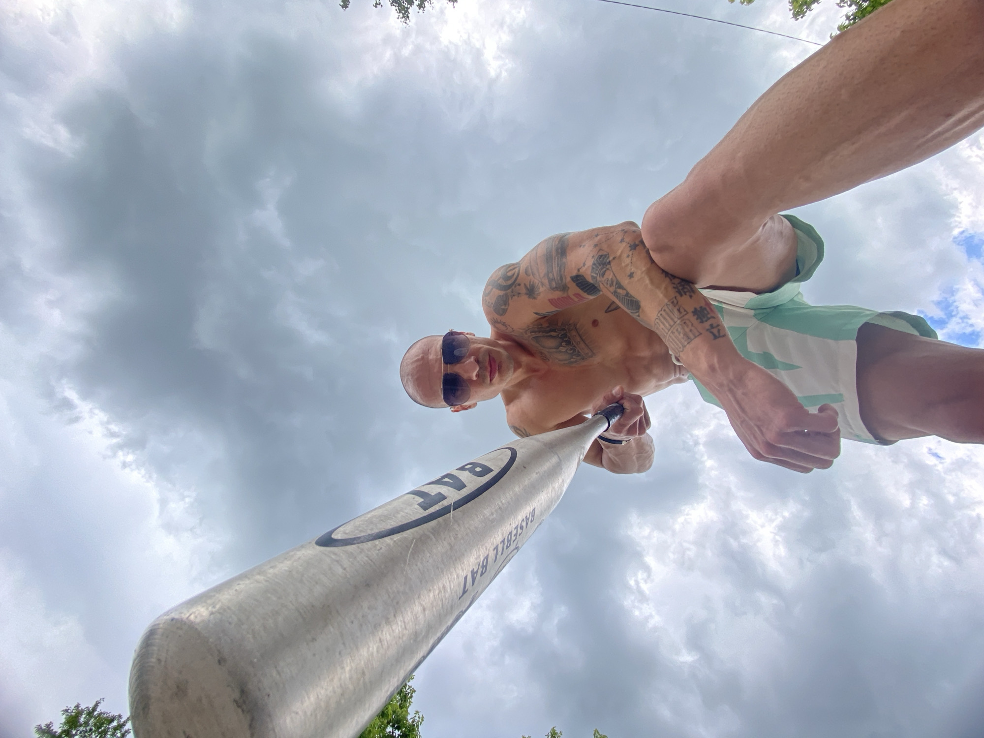 A low-angle shot depicts a man holding a baseball bat. He is bald, wearing sunglasses, and has numerous tattoos on his arms and chest. He's wearing light green and white shorts. The baseball bat is pointed towards the camera. The sky is overcast with dark clouds. The specific location in Pleven, Bulgaria, is not discernible from the image.