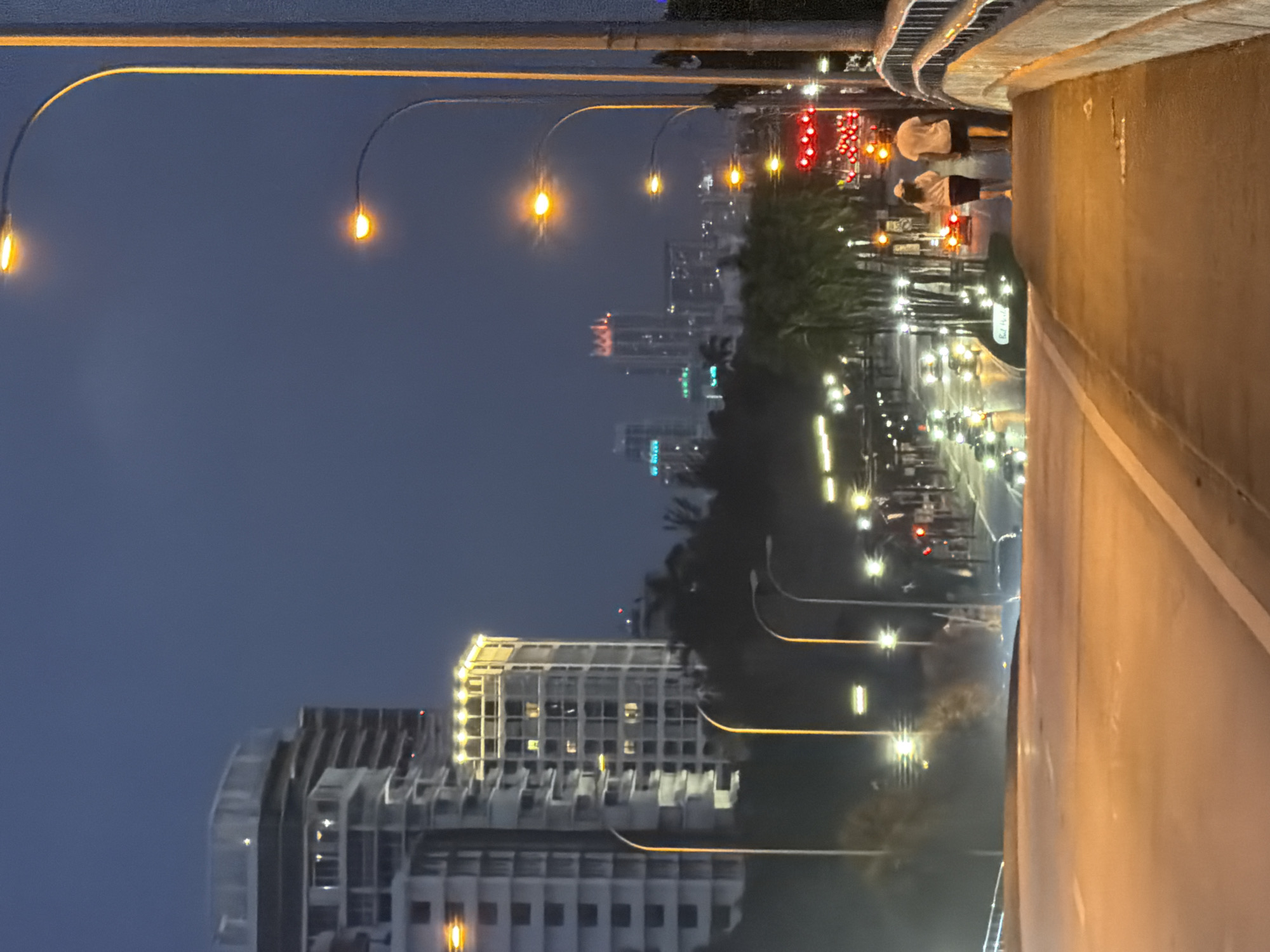 This is a photograph taken at dusk or night on a bridge in Bal Harbour Village, United States of America, overlooking a cityscape and a busy street. The foreground features a concrete bridge with a walkway. Two individuals, a man and a woman, are walking away from the camera on the walkway. The man is wearing a white t-shirt and shorts, and the woman is wearing a white cap, a white top, and a dark skirt.
In the mid-ground, there is a street with multiple lanes of traffic, illuminated by streetlights and the headlights and taillights of vehicles. A traffic light showing red is visible. Palm trees line the street. In the background, a city skyline is visible with several tall buildings, some of which are illuminated. One prominent building appears to be under construction. The sky is dark blue, indicating nighttime. The overall atmosphere is urban and illuminated.