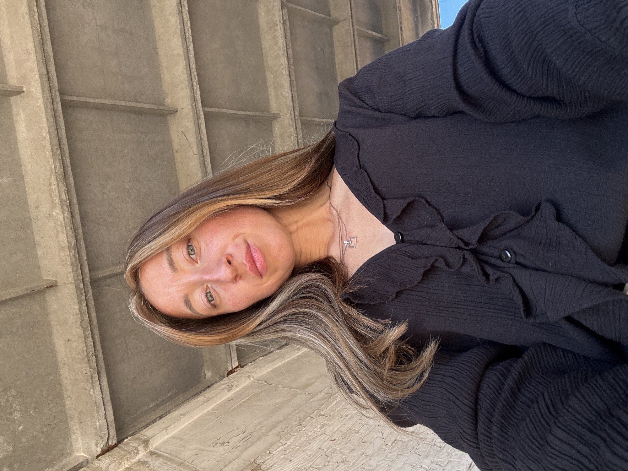 A woman with long, wavy brown and blonde hair is looking up at the camera. She is wearing a black blouse with ruffled details around the neckline and buttons down the front. A silver necklace with a square pendant is visible around her neck. She is positioned under a concrete structure, possibly a bridge or overpass, with visible beams and a textured ceiling. To her left, a brick wall is visible. The background appears to be outdoors with a hint of blue sky in the upper right corner. The image is a selfie, taken from a low angle. The woman's face is illuminated by natural light, highlighting her green eyes and fair skin with freckles. The overall scene suggests an urban environment, but the specific location in Chaikovsky, Russia cannot be determined from the visual information alone.