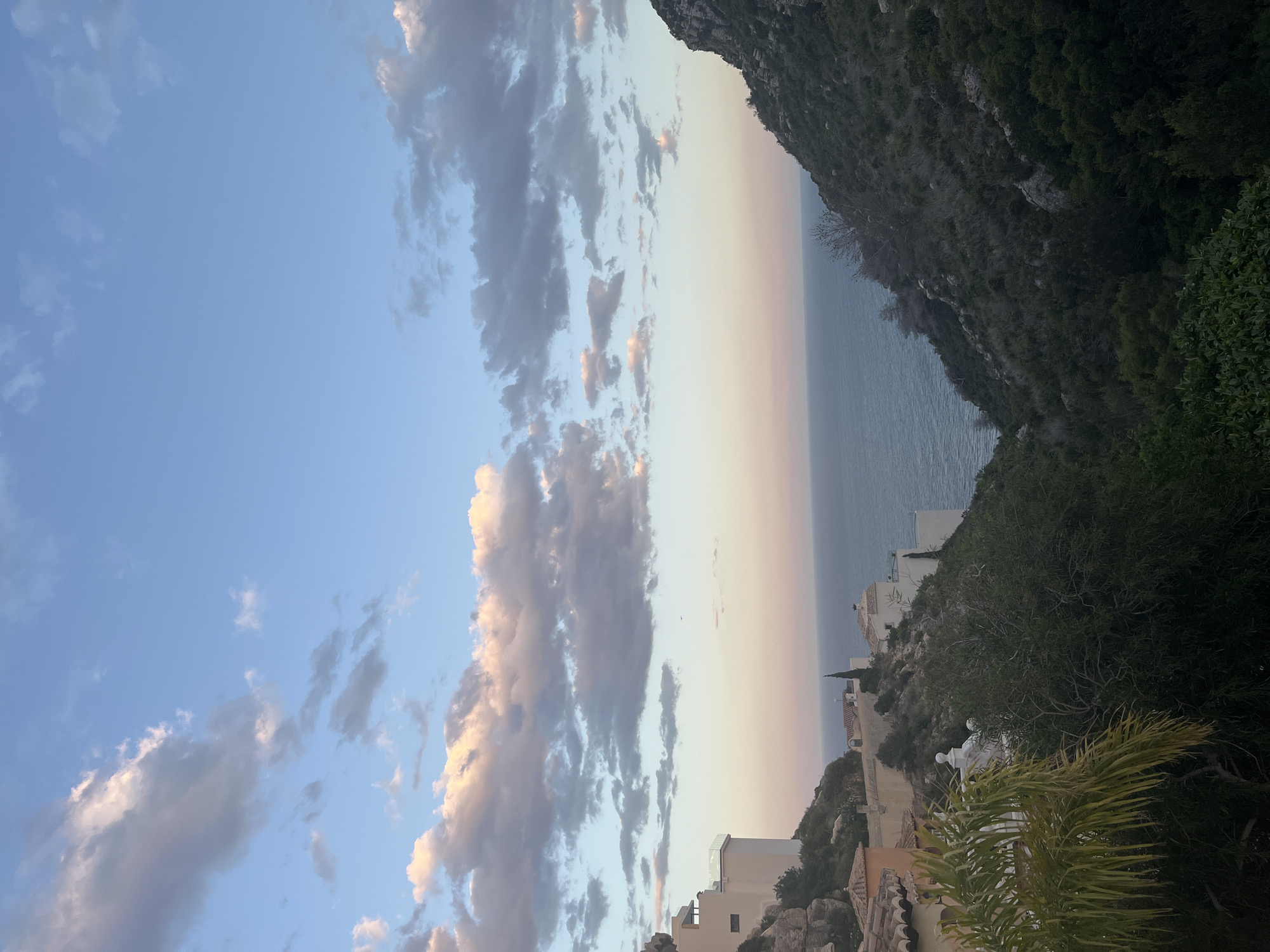 The image shows a coastal view in El Poble Nou de Benitatxell / Benitachell, Spain, during what appears to be late afternoon or early evening. The sky is predominantly blue, with streaks of grey and pale pink clouds. The horizon displays a gradient of colors, ranging from pale pink to light blue where the sky meets the sea.
The sea is a dark blue color. The foreground features a rocky, mountainous coastline covered in sparse vegetation and clusters of trees. Buildings are visible nestled among the terrain.
There are no people visible in the frame. The focus is on the natural landscape, architectural elements, and atmospheric conditions.