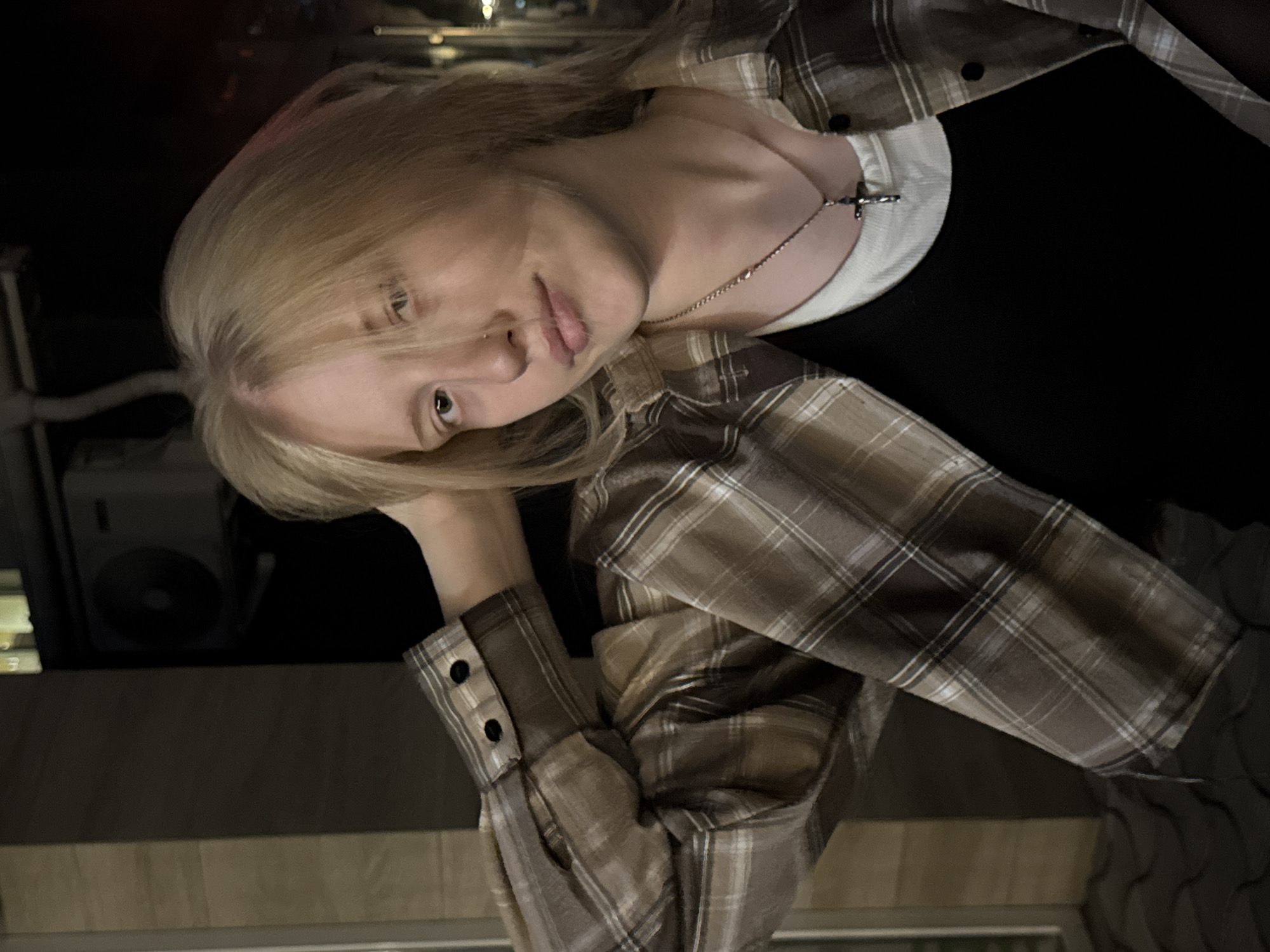 A young person with blonde hair, wearing a plaid shirt over a black tank top and a white undershirt, poses with their hand behind their head. A silver cross necklace is visible. The background suggests an urban setting, possibly Tiraspol, Moldova, with architectural elements and an air conditioning unit visible on a building. The lighting indicates it is either evening or night.