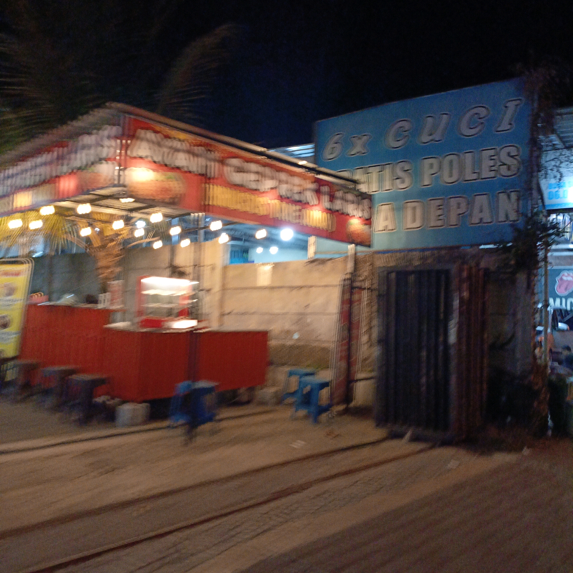 The image depicts an outdoor food stall at night, possibly in Salatiga, Indonesia. A bright red structure with a corrugated metal roof is visible, illuminated by numerous string lights. Signs with text are present, including one in blue with white lettering that appears to say "6x CUCI GRATIS POLES A DEPAN". Another sign, red with yellow and white lettering, is partially obscured and appears to advertise food. In front of the stall, there are blue plastic stools and a red counter. The ground appears to be dirt or gravel, with what looks like railway tracks running through it. The overall scene suggests a bustling night market or roadside eatery.