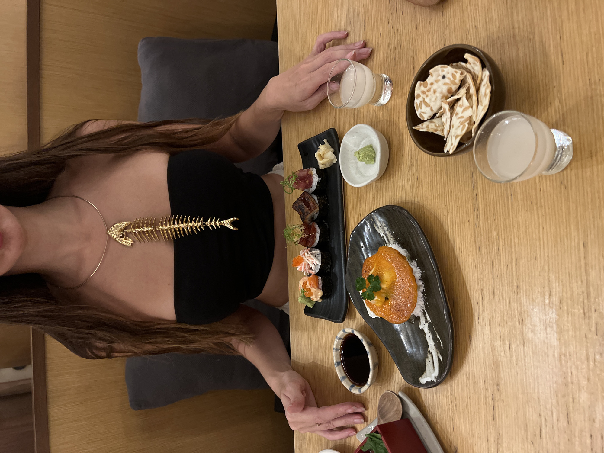 A young woman with long brown hair and wearing a black strapless top and gold fishbone necklace is seated at a light wooden table. The table is set with various Japanese dishes, including a plate of sushi with wasabi and ginger, a black plate with a singular fried dish decorated with a white sauce and parsley, a bowl of soy sauce, a bowl of crackers, and two glasses of milky sake. The woman has her hands on the table, one near a glass of sake and the other near the soy sauce bowl. The scene is likely taking place at a restaurant in North Kuta, Indonesia.