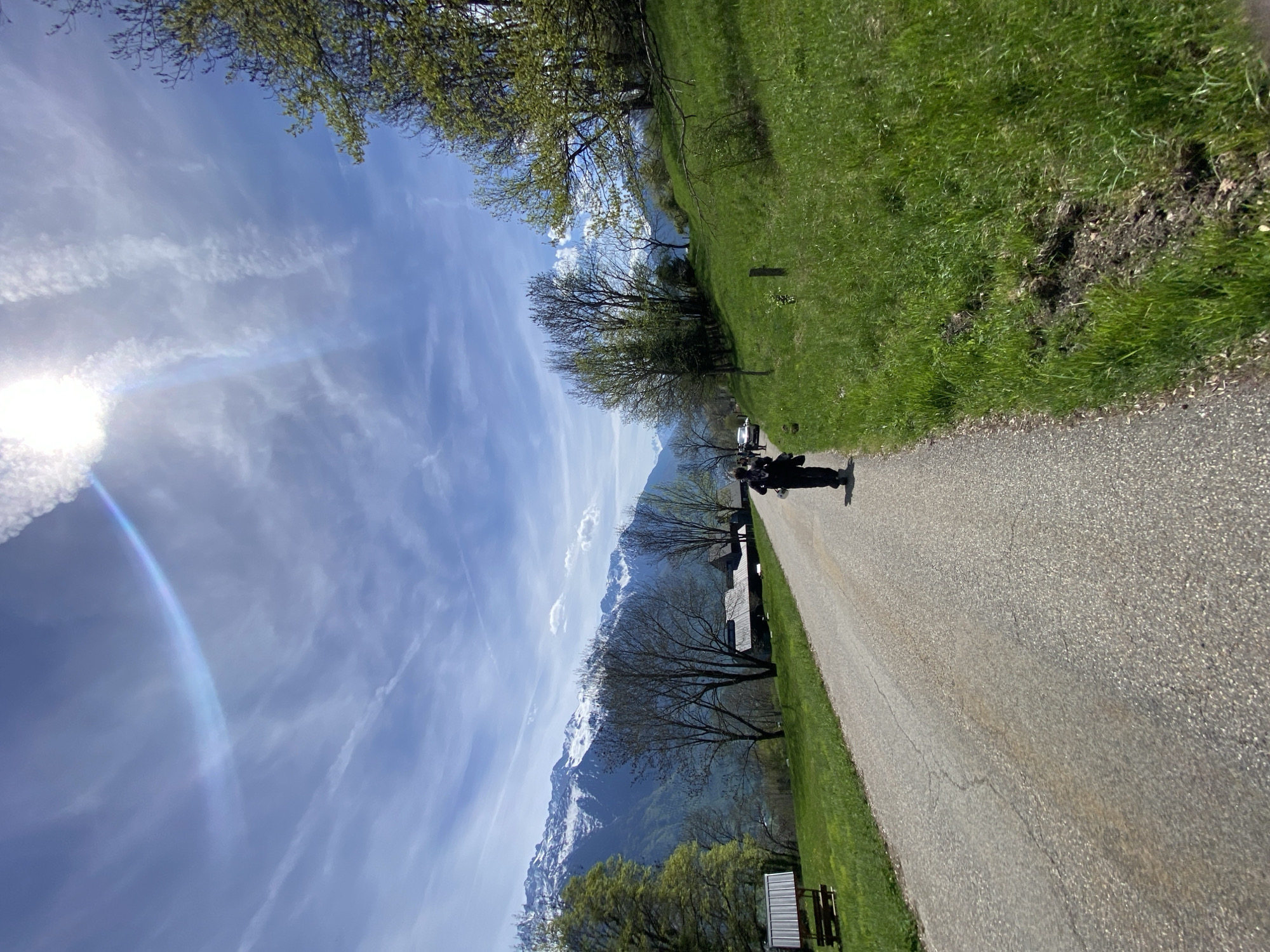 This image captures a vibrant, sunny mountainous landscape, likely during spring, in Saint-Georges-d'Hurtières, France. The scene is dominated by a clear blue sky filled with wispy white clouds and distinct contrails. The sun, prominent in the upper left, casts a bright glare and lens flare, including a subtle rainbow-like arc.
In the middle ground, majestic snow-capped mountains stretch across the horizon, their lower slopes covered in dark forests, indicating a high-altitude setting. A paved asphalt path, slightly curving, extends from the lower right foreground towards the distant mountains. Lush green grass borders both sides of this path.
On the left side of the path, several trees are visible, some already in full green foliage, while others still show bare or budding branches, reinforcing the early spring season. Nestled among these trees and further in the distance are a few indistinct buildings or structures. In the lower left, a wooden bench or similar low structure is partially visible.
A single person, dressed in dark clothing, stands on the right side of the path, facing towards the mountains, their figure silhouetted against the bright background. Their activity appears to be observing the scenery or taking a leisurely walk. A small, vertical wooden post is also visible on the grassy verge to the right.
The bright sunlight suggests it is midday or early afternoon. The overall impression is one of natural beauty and tranquility in a scenic rural or mountainous environment. No discernible text is visible within the image.