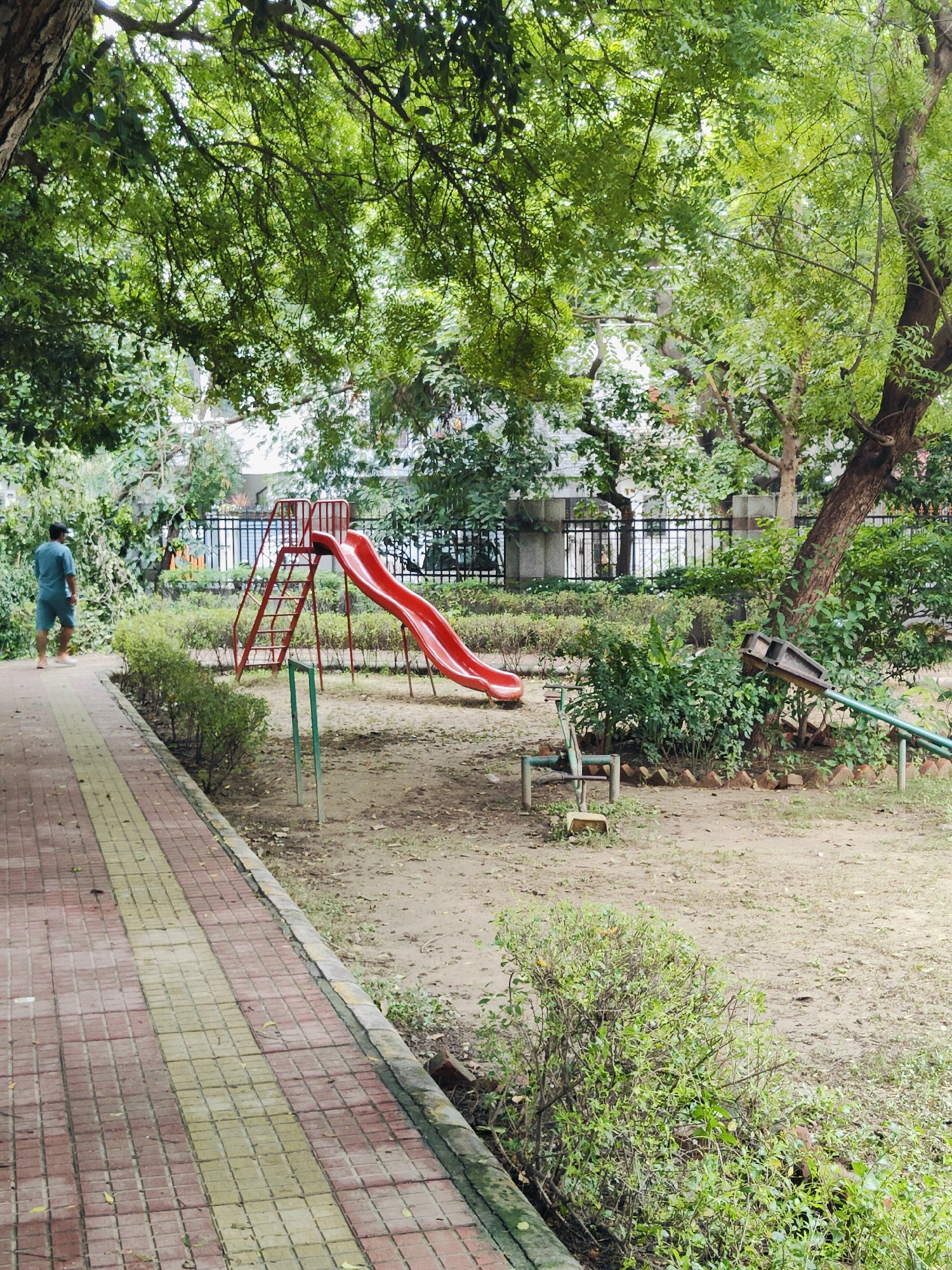 This is a medium-shot, eye-level, outdoor photograph of a children's playground in Gurgaon, India. The scene is dominated by lush green foliage and trees, with a red metal slide and a green metal seesaw visible in the center of the frame. A person, wearing a blue shirt and shorts, walks away from the camera on a red and yellow tiled pathway on the left side of the image. The ground is covered in dirt and dry leaves. A black metal fence runs along the background, behind which a building is partially visible. The overall atmosphere is peaceful and serene.