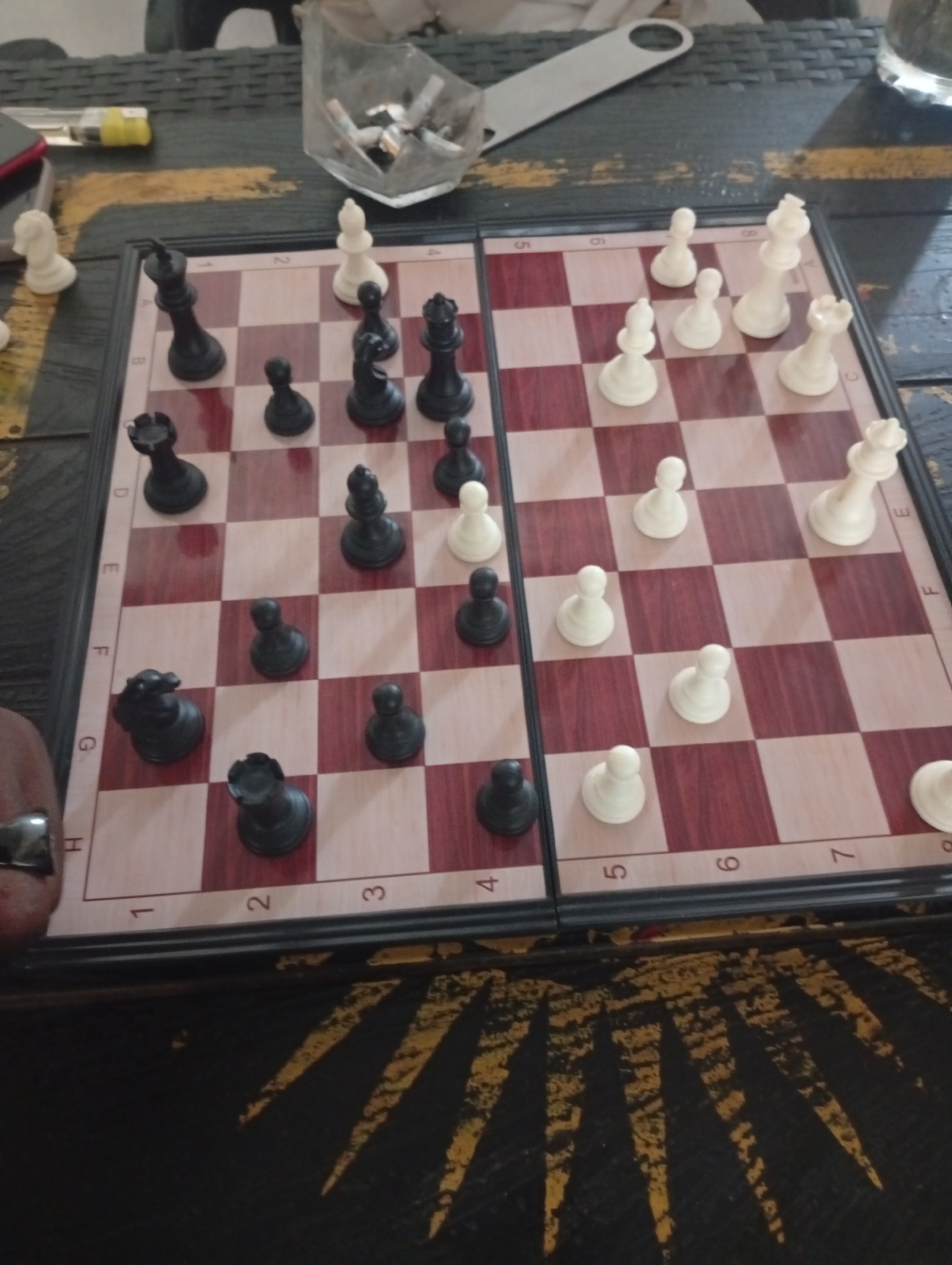 The image captures a chessboard set up on a dark, patterned table, likely indoors in Barakin Akawo, Nigeria. The chessboard is foldable, indicated by the seam down the middle, and features alternating light and dark red squares. Both black and white plastic chess pieces are scattered across the board, suggesting a game is either in progress, just finished, or about to begin, as they are not in their initial setup positions.
On the left side of the board, a human hand with a silver ring is partially visible. Surrounding the chessboard are various items: a clear, glass-like container functioning as an ashtray filled with cigarette butts, a silver bottle opener, and a yellow lighter. A corner of what appears to be a red mobile phone is also visible. The table surface is dark, possibly black, with worn yellow patterns, giving it a somewhat rustic or well-used appearance.
Visible alphanumeric text on the chessboard includes ranks '1', '2', '3', '4', '5', '6', '7' along the sides, and files 'H', 'G', 'F', 'E', 'D', 'C'. Some numbers like '5', '6', and '2' are also visible near the white pieces, and '5', '6', '7' near the black pieces, likely representing ranks. The overall scene depicts a casual environment for a game of chess.
