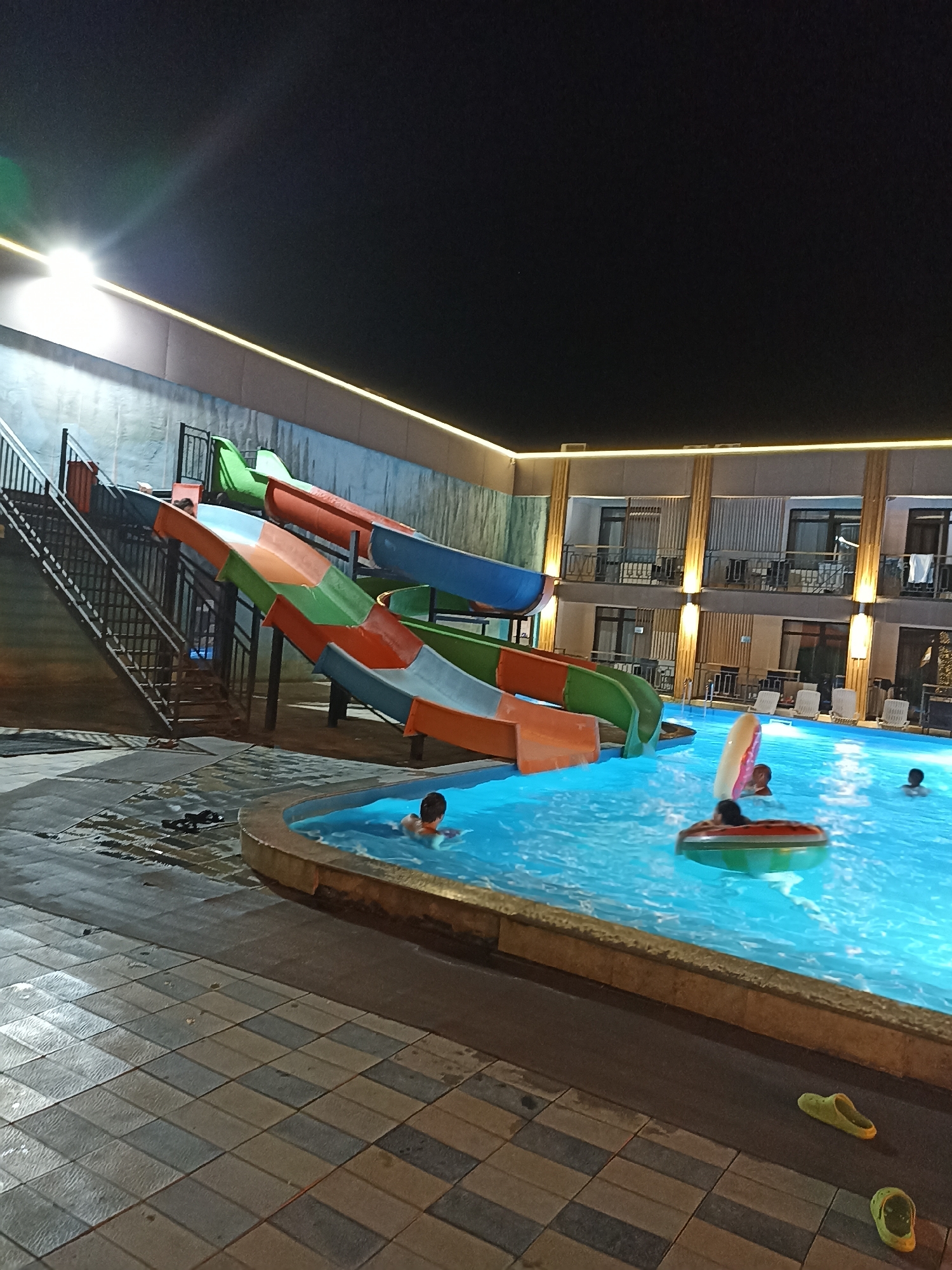 The image depicts an outdoor swimming pool area at night, likely located in Sorbulak, Kazakhstan. A multi-colored water slide structure dominates the left side of the frame, with green, orange, and blue sections. To the right of the slide, a swimming pool filled with brightly lit blue water is visible. Several individuals are in the pool; one person is visible in the foreground, partially submerged and facing away from the camera. Two inflatable rings are also present in the water. The surrounding area features a tiled walkway and the facade of a building with multiple stories, balconies, and illuminated windows. Yellow accent lighting runs along the top of the building. The overall scene suggests a recreational facility operating after dark.