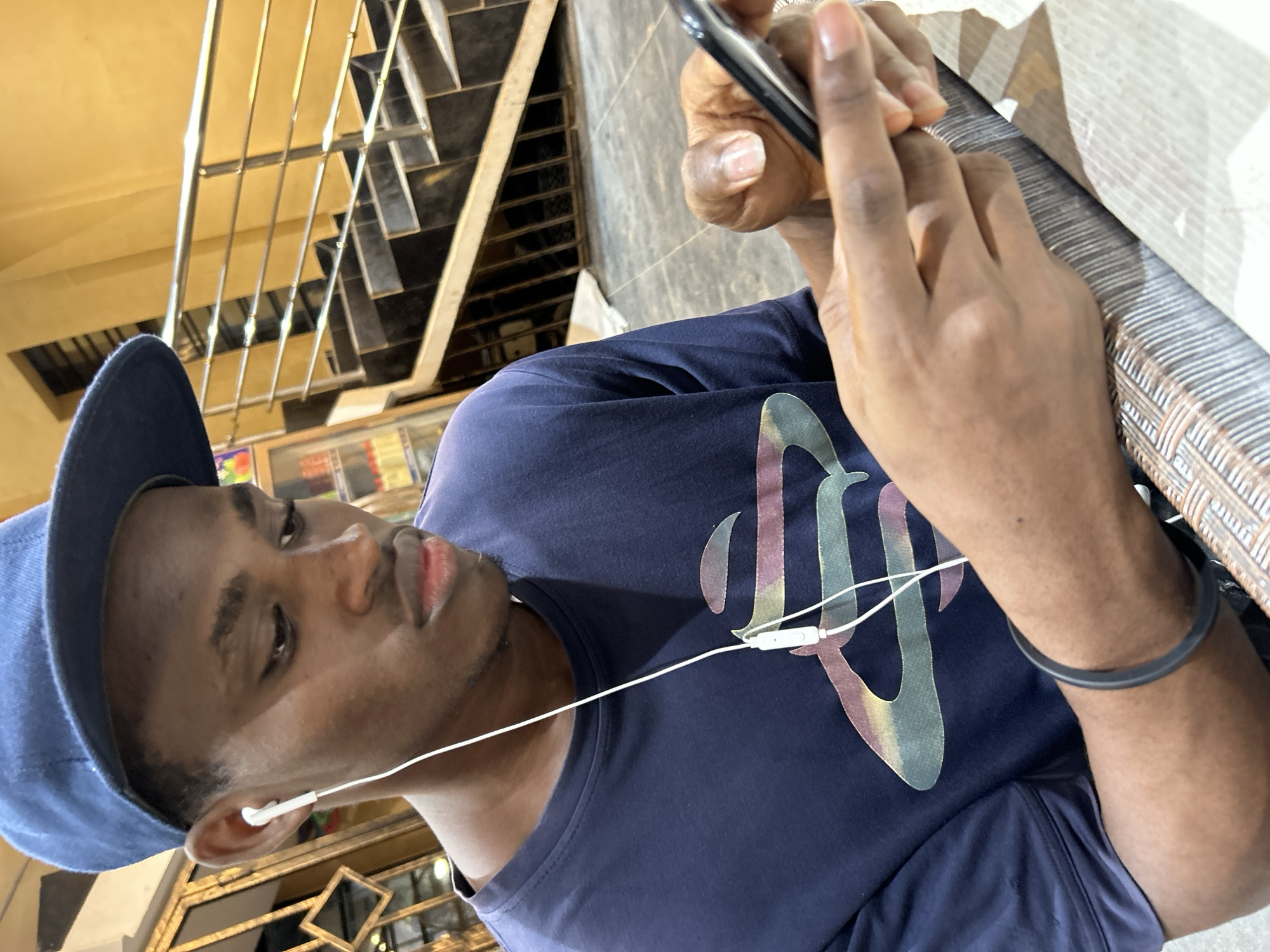 A young Black male wearing a blue baseball cap and a dark blue t-shirt with a stylized logo is shown from the chest up, looking down at a smartphone held in his hands. He has white earbuds in his ears, with the cord visible. He is wearing a black wristband on his left wrist. The background shows a staircase with a silver railing, and what appears to be a shop or building interior with shelves of products visible behind him. The scene is likely indoors, possibly in a public space such as a cafe or shop, in Ihiagwa, Nigeria.