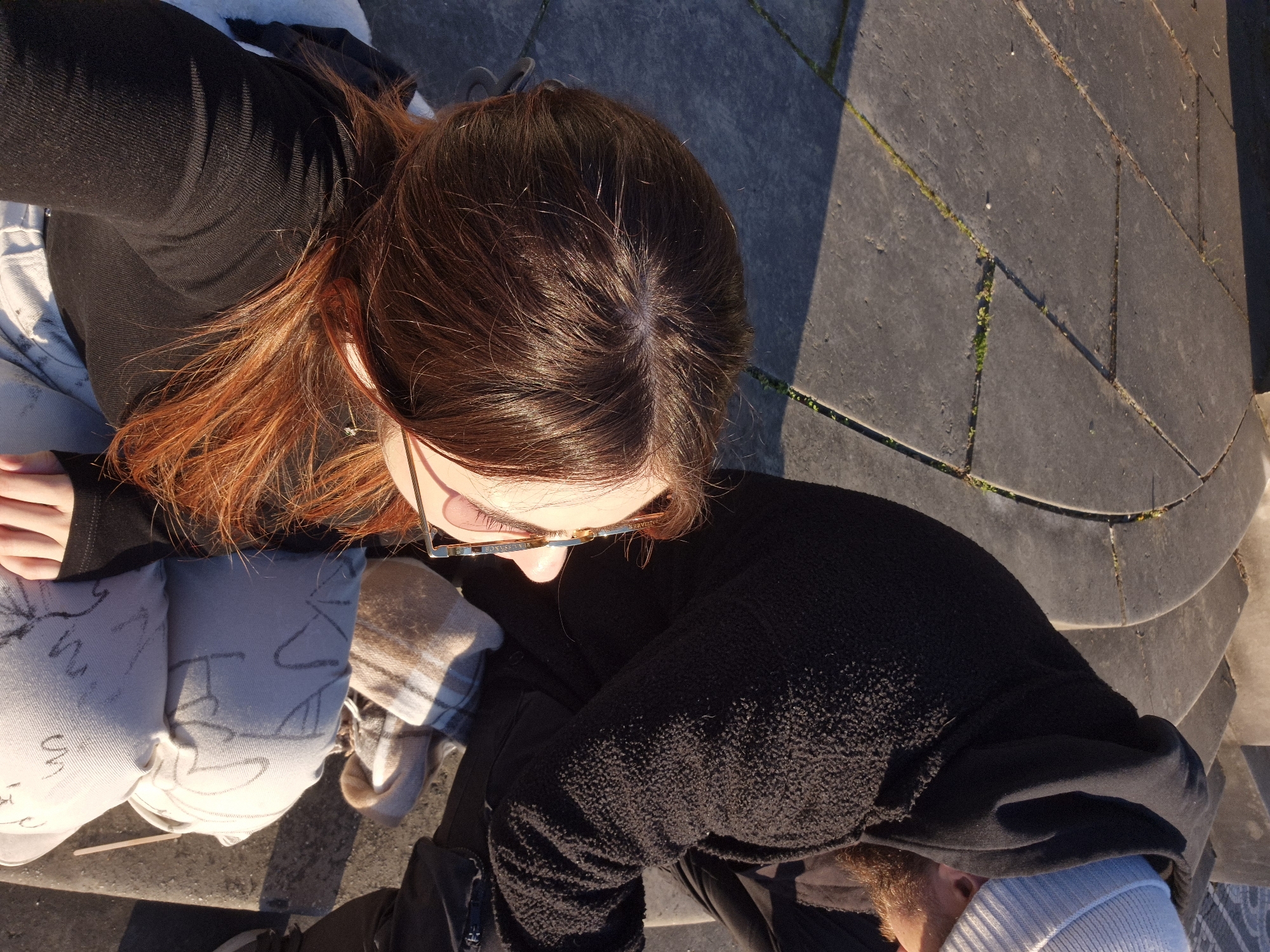 The image depicts two individuals seated outdoors on grey stone steps or pavement, likely in a public area of Strasbourg, France, under direct sunlight.
In the foreground, the upper back and right side profile of a person are visible. This individual has reddish-brown hair, is wearing a black long-sleeved top, and has sunglasses positioned on their face. They are resting on light-colored, possibly off-white, fabric or cushions which feature faint dark, abstract markings. Their right hand is visible resting on the fabric.
To the right and slightly behind, a second individual is seen from the side and back. This person has a visible beard, wears a light grey knitted beanie, and a dark, textured hooded jacket, possibly fleece.
Between the two individuals, a folded plaid-patterned fabric, resembling a scarf or blanket, is visible. The ground consists of large, irregular grey paving stones with visible seams, some containing small patches of green vegetation. Strong sunlight illuminates the scene from the right, casting distinct shadows on the ground and structures. The individuals appear to be resting or observing the environment.