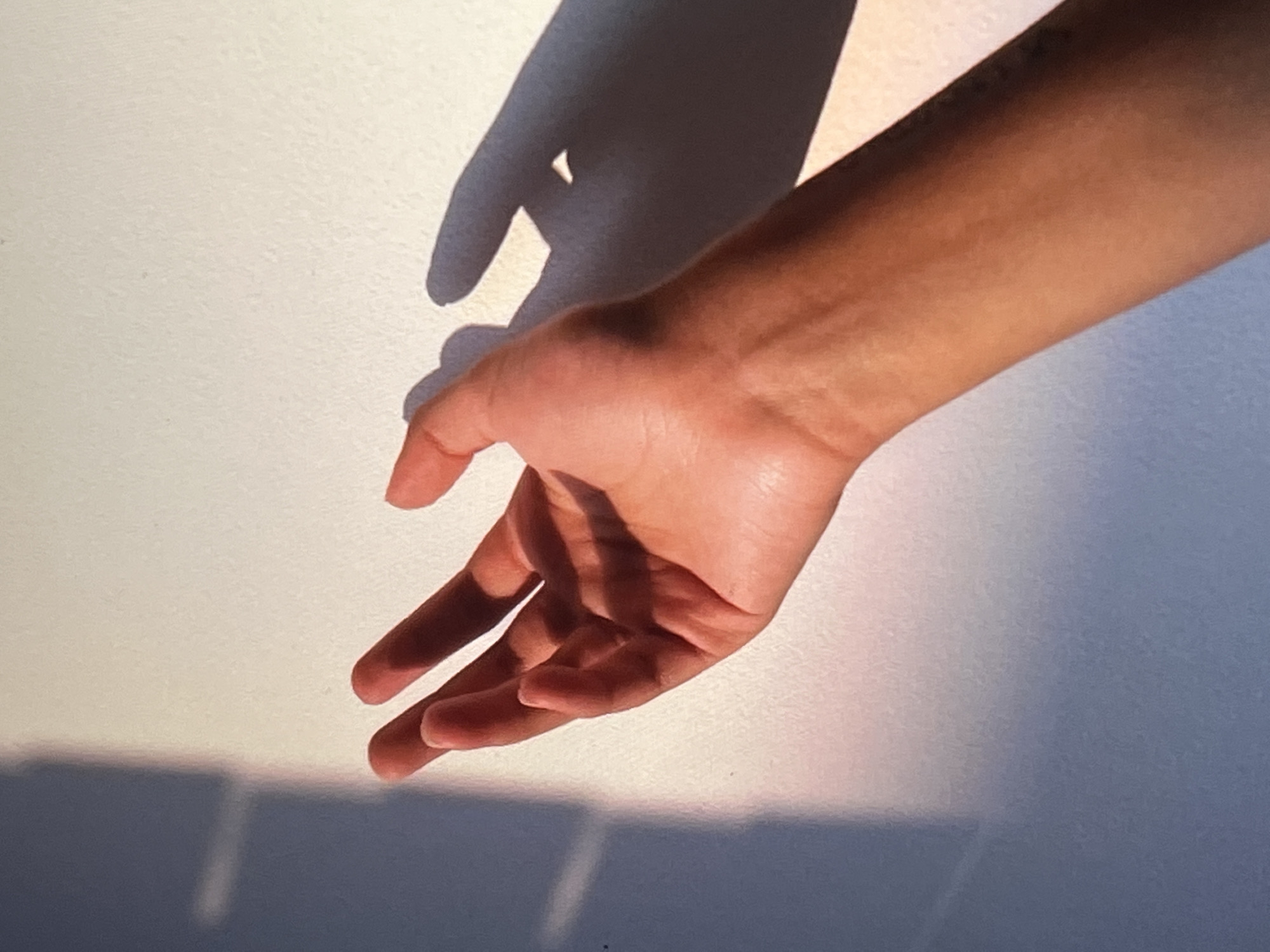 A close-up, eye-level shot features a person's right arm and hand extended toward a white wall bathed in warm, angled sunlight, casting a distinct shadow. The hand is open, palm facing away from the viewer, with fingers slightly curled. The skin tone of the arm and hand appears to be a warm, tanned hue. The wall exhibits subtle texture and is partially illuminated by the sunlight, creating a gradient of light and shadow. On the left side of the image, a darker shadow falls across the wall, originating from an unseen object. A faint pinkish glow is visible at the edge of the shadow on the wall, suggesting the quality of the light. The background is the white wall, with some slight variations in tone due to the lighting. No specific location in Genemuiden, Netherlands, can be identified from the provided image.
